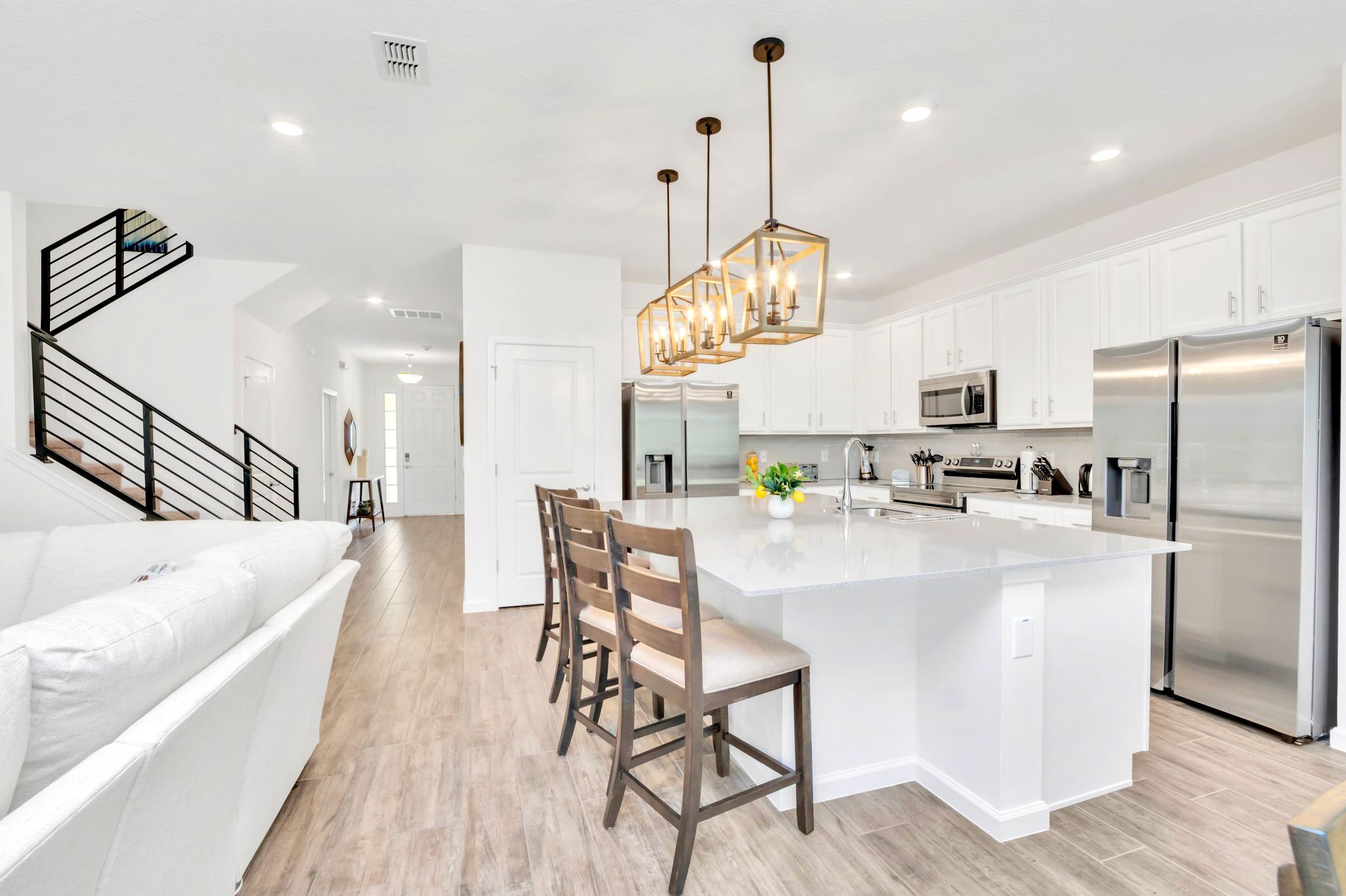 A modern kitchen featuring a large white island with a sink and bar seating, illuminated by three industrial-style pendant lights, with stainless steel appliances visible in the background.