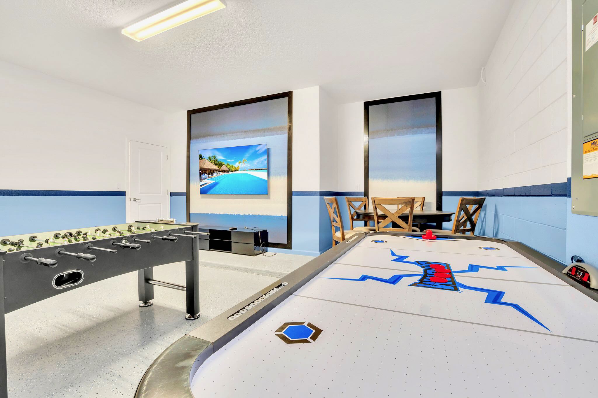 A closer view of the game room emphasizes the air hockey table with blue accents and the large wall-mounted TV, set against a white and blue painted wall, with a foosball table partially visible.