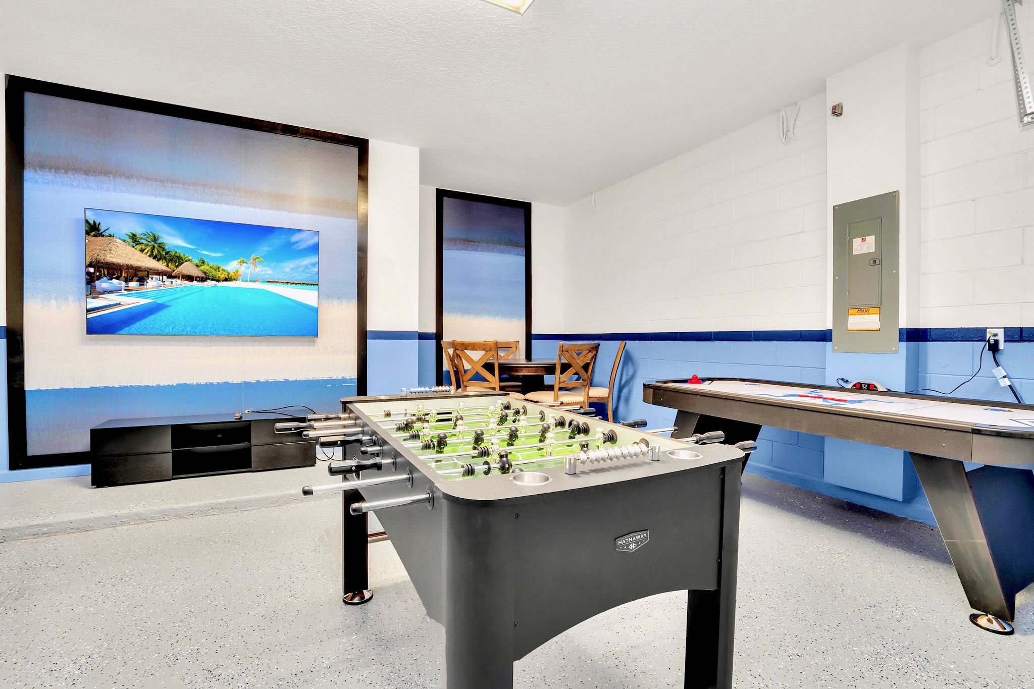 A vibrant game room features a foosball table and an air hockey table on a speckled gray floor, with a large wall-mounted TV displaying a beach scene and a small dining set in the background, all within a white and blue painted space.