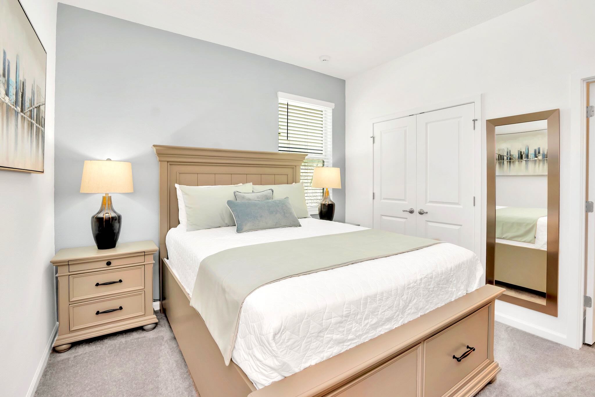 A bright bedroom featuring a light brown wooden bed frame with white and light blue bedding, flanked by two matching nightstands with lamps, and an accent light blue wall behind the bed.