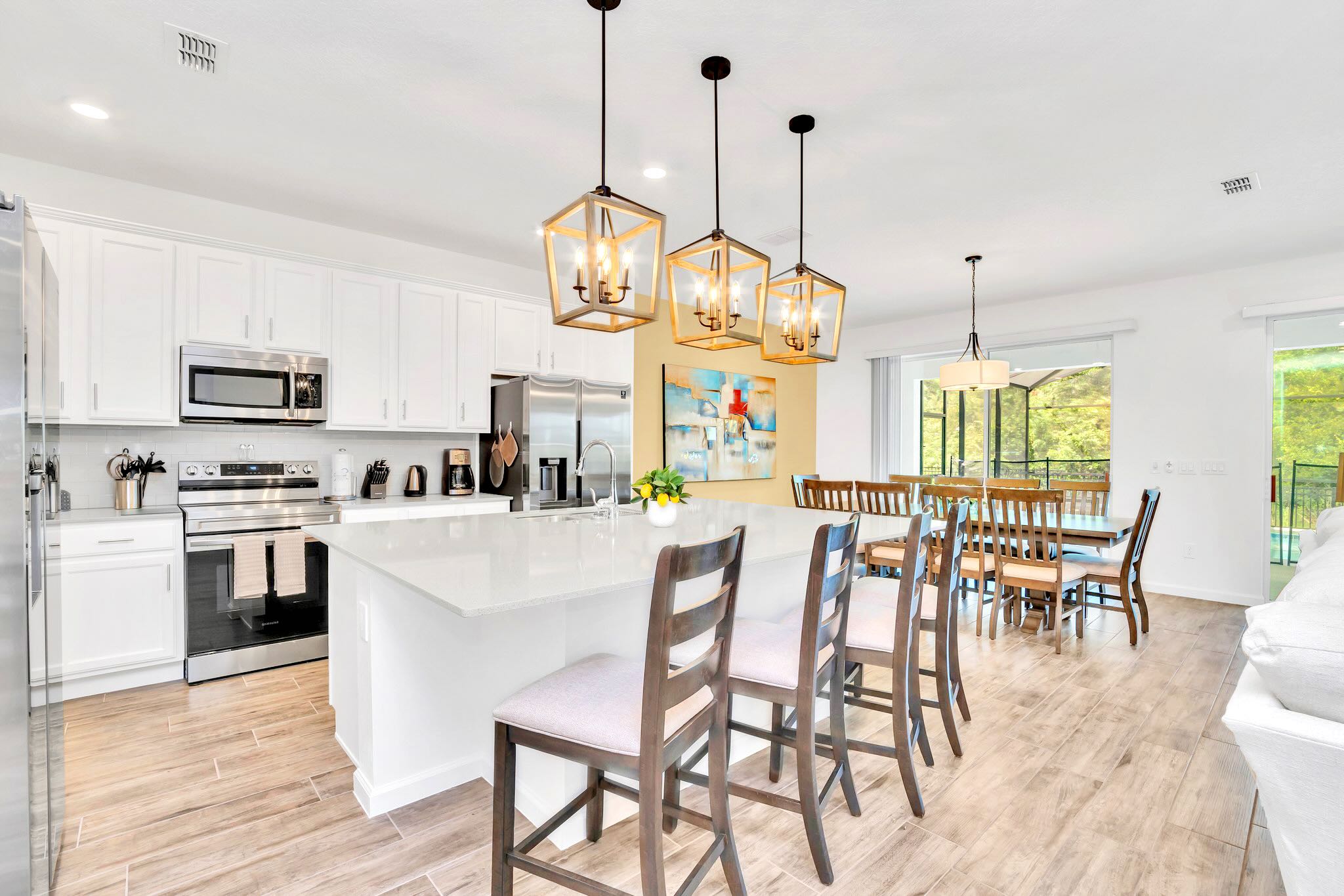A bright, modern kitchen features white shaker cabinets, stainless steel appliances, a large island with white quartz countertops and four bar stools, and three industrial-style pendant lights, opening to a dining area with a long wooden table.