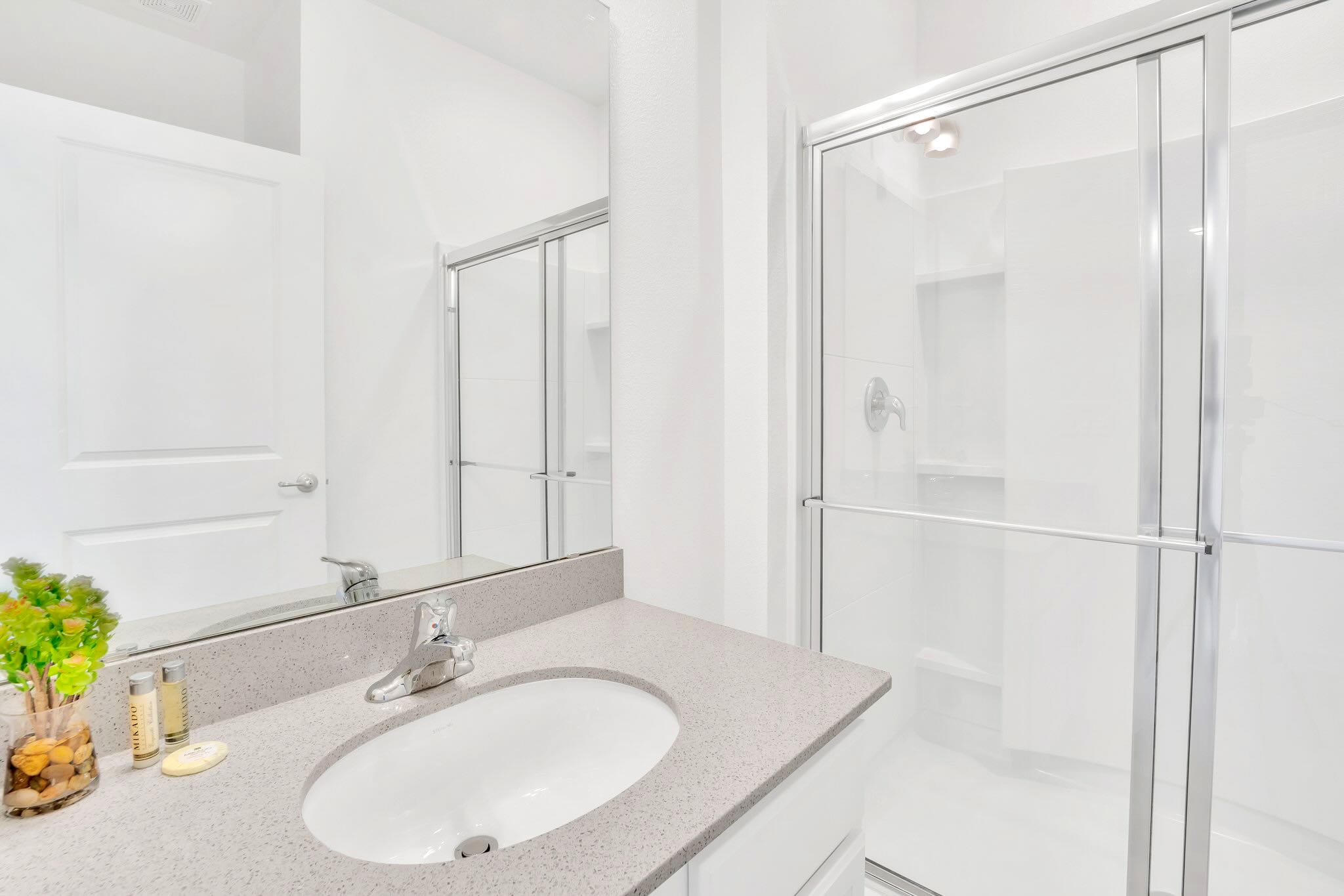 A bright bathroom featuring a single vanity with a grey speckled countertop and a large mirror, alongside a modern walk-in shower with a clear glass enclosure.