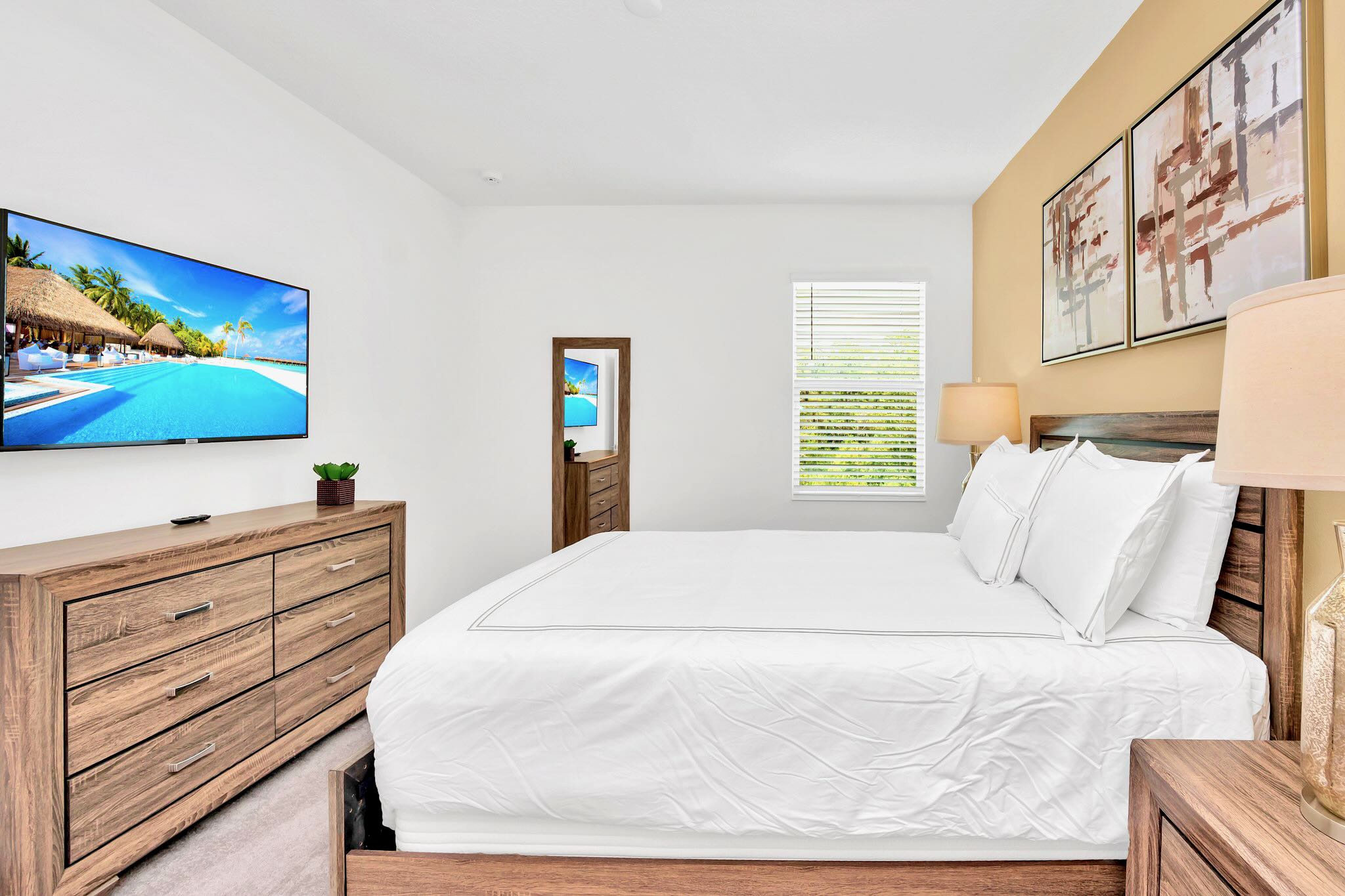 A modern bedroom with a dark wood bed frame, white bedding, a large flat-screen TV mounted above a matching dresser, and a warm yellow accent wall with abstract art.