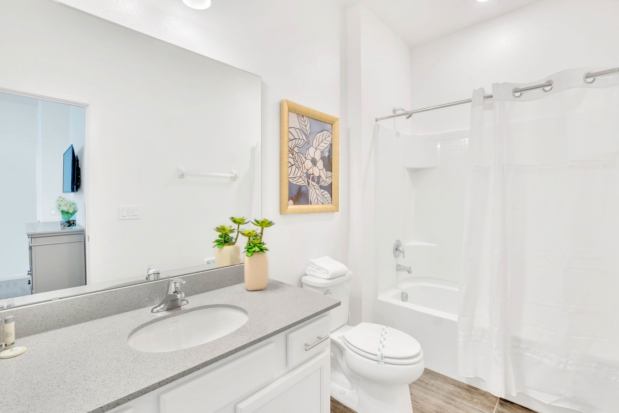 A clean white bathroom with a single vanity featuring a grey speckled countertop, a large mirror, a toilet, and a bathtub with a white shower curtain.
