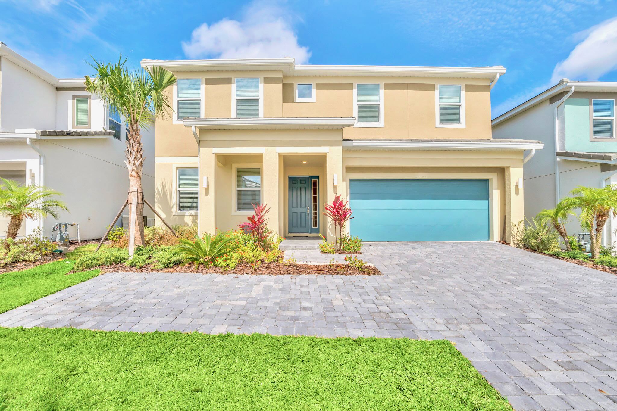 The front exterior of a two-story house features light beige stucco, a vibrant blue front door, and a matching blue garage door, with a paver driveway, green lawn, and tropical landscaping under a bright blue sky.