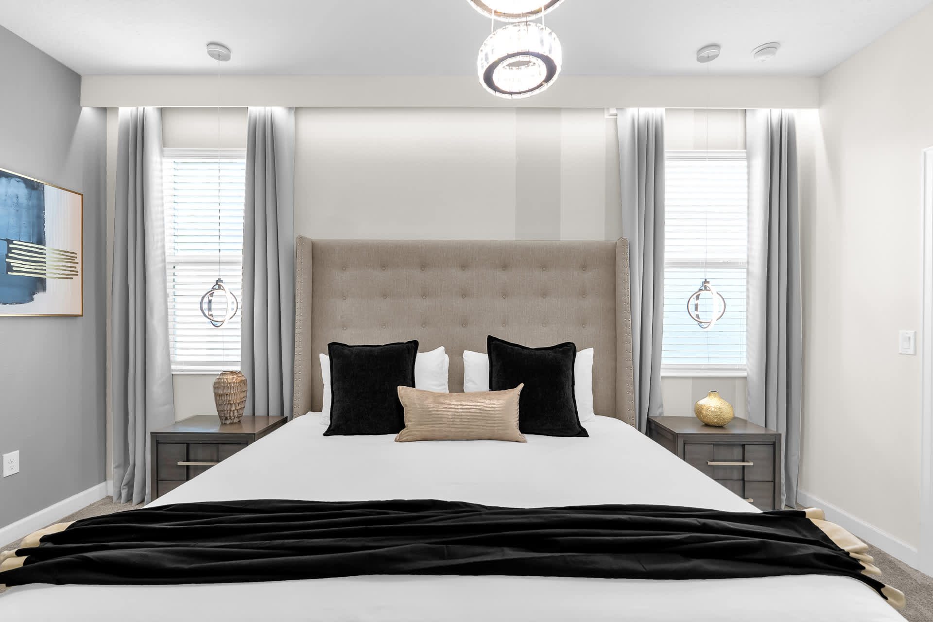 A close-up view of a king-size bed with a tall upholstered headboard, black and gold decorative pillows, and two nightstands with unique hanging pendant lights. Large windows with blinds and curtains flank the bed.
