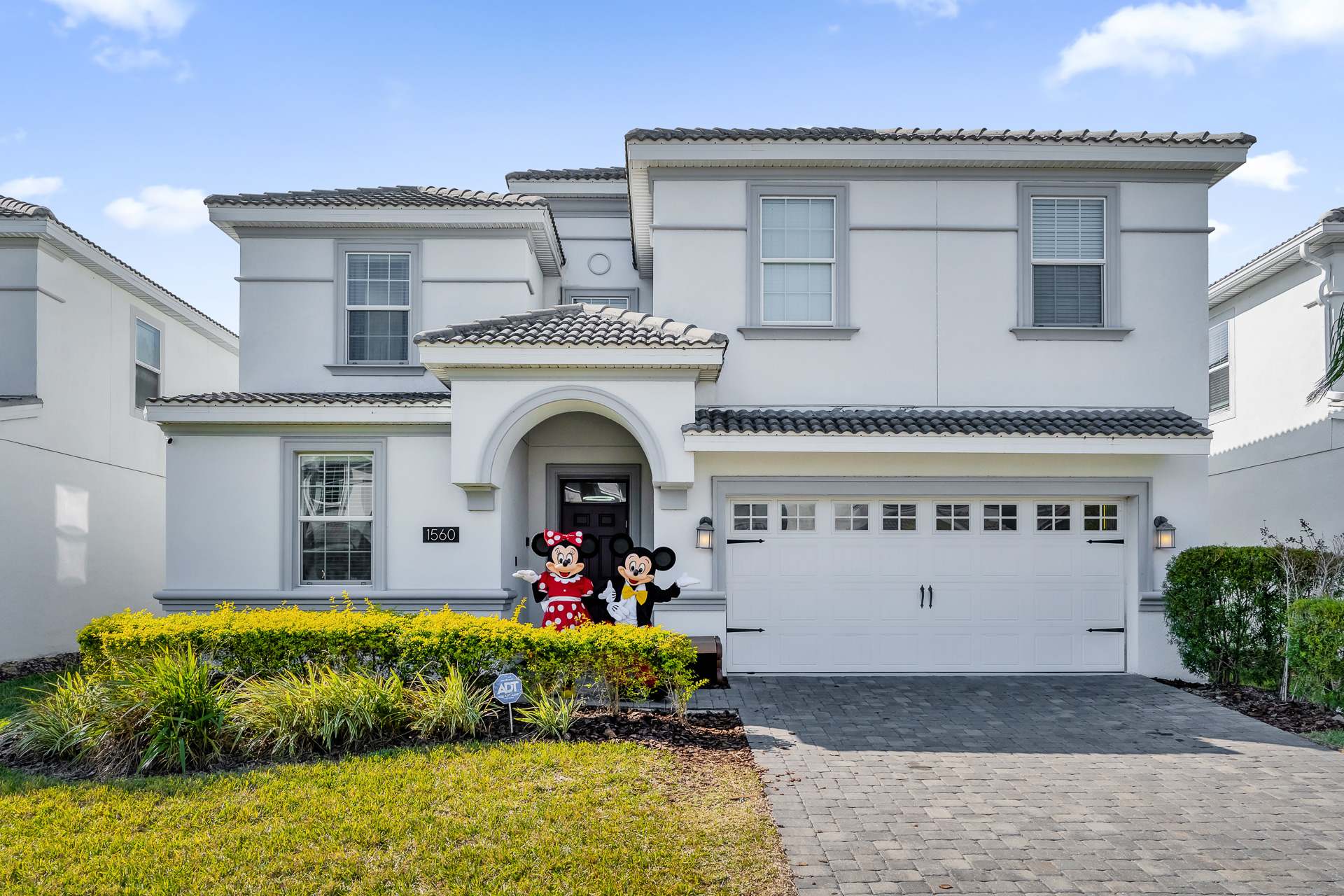Front exterior of a modern two-story white house with gray trim, a gray tiled roof, and a paved driveway leading to a two-car garage. Mickey and Minnie Mouse statues stand by the arched front entrance.