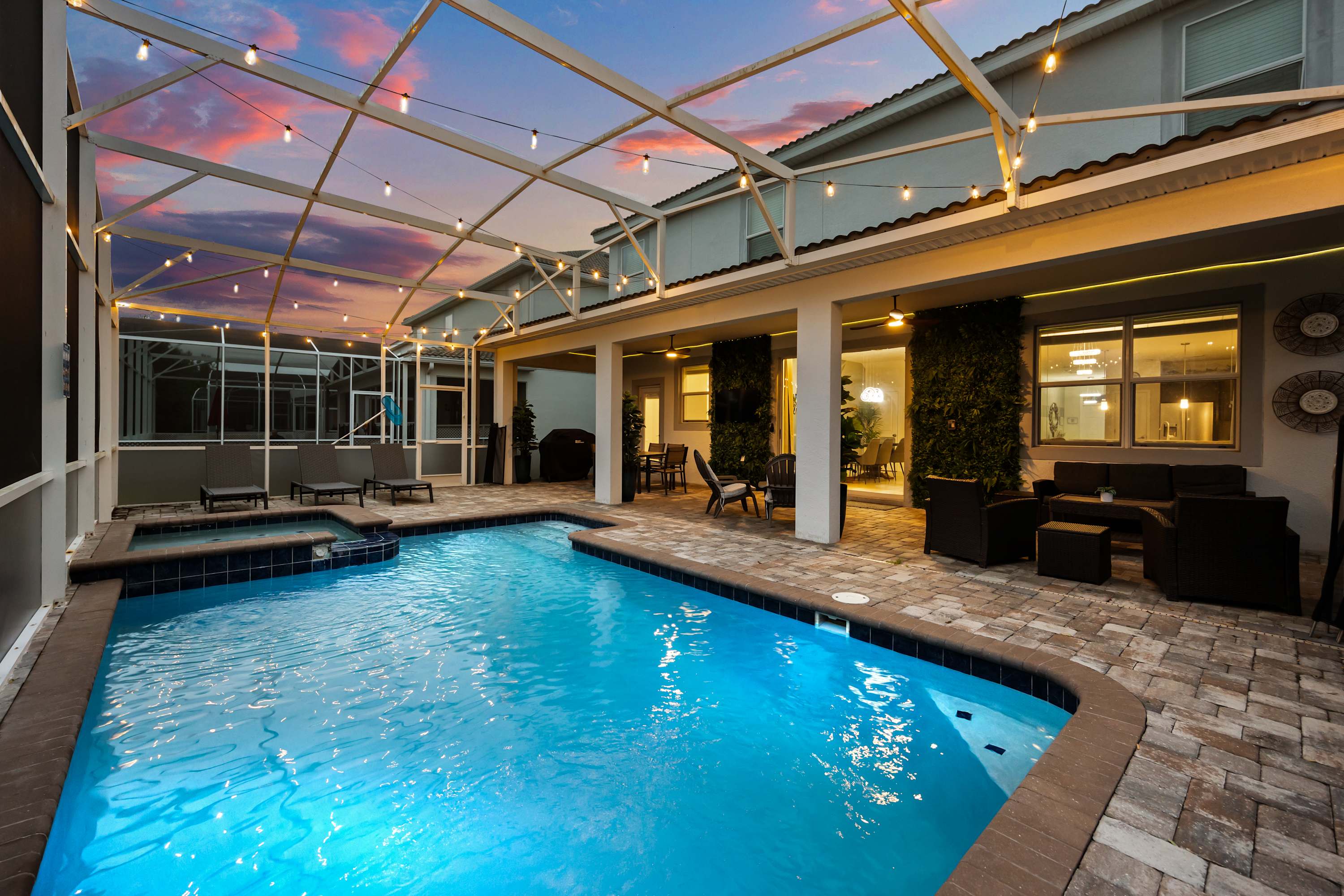 Spacious screened-in pool area featuring a rectangular private pool, an integrated hot tub, and a paved patio with outdoor seating, all under string lights at sunset.