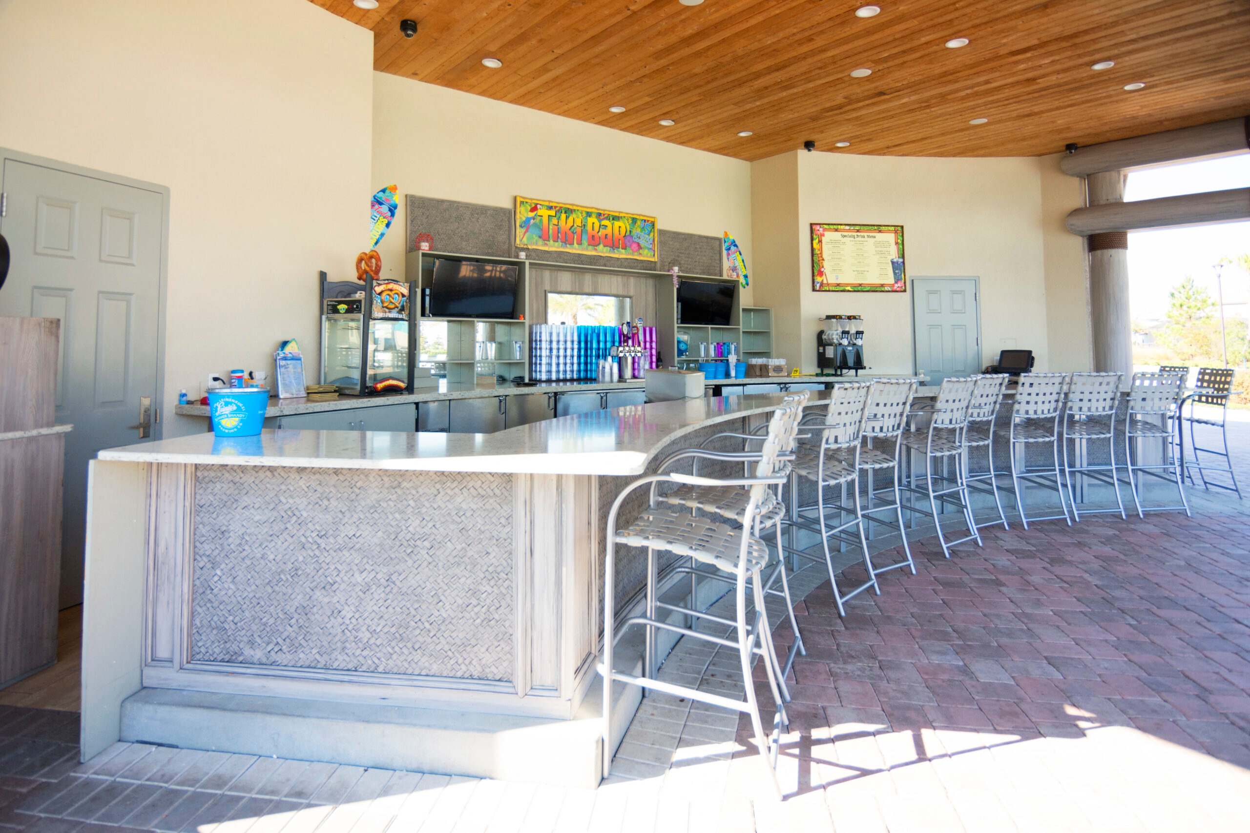 An inviting outdoor tiki bar with a long, curved counter and numerous bar stools, featuring a warm wooden ceiling and a brick-patterned floor. Various drinks, bar equipment, and two flat-screen TVs are visible behind the counter.