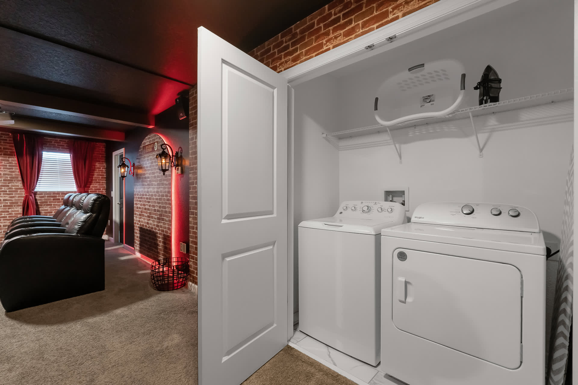 A compact laundry closet features a white top-loading washing machine and a matching dryer, with a wire shelf above holding a laundry basket and an iron. To the left, a dark media room with brick-patterned walls and recliner seating is visible.