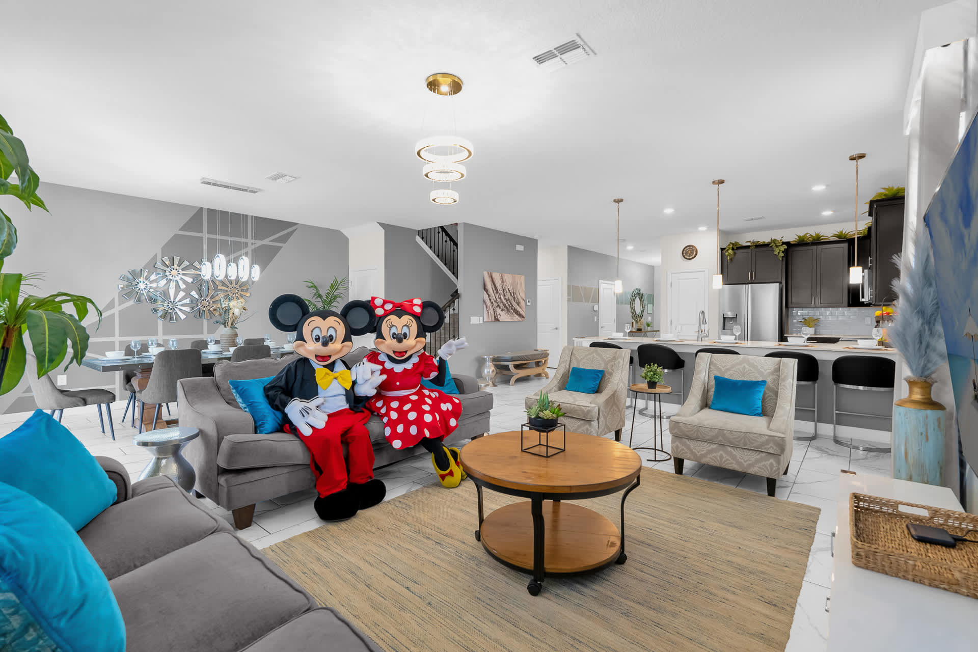 A spacious living room featuring a gray sofa with blue pillows, a round wooden coffee table, and two armchairs. Mickey and Minnie Mouse characters are seated on the sofa, with the dining area and kitchen visible in the background.