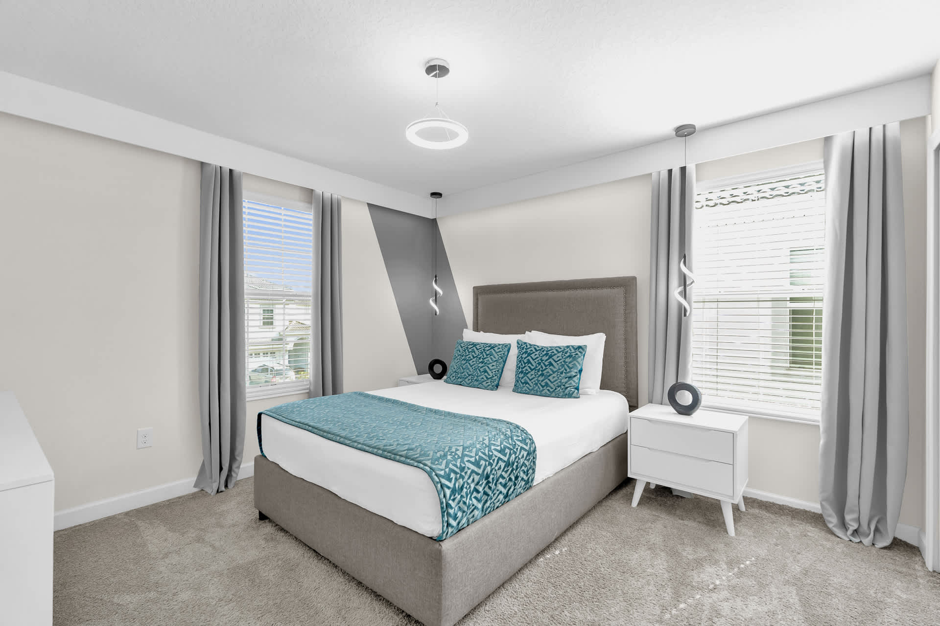 Bright bedroom with a gray upholstered queen bed, blue accent pillows, and a teal patterned throw. The room features a gray and white diagonal accent wall, a circular ceiling light, and two white nightstands with unique pendant lights.