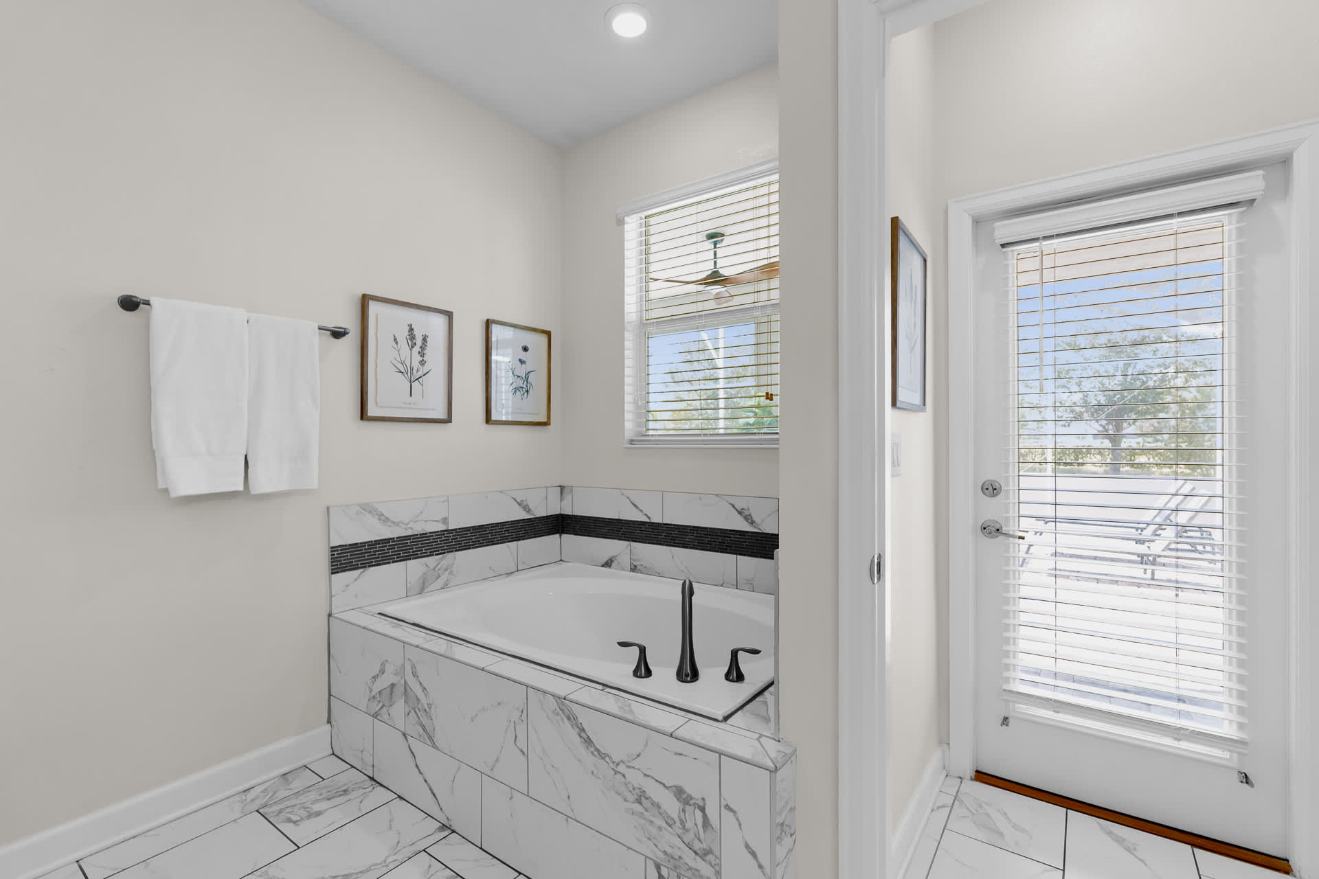 Spacious bathroom with a built-in soaking tub surrounded by white marble-patterned tiles and a dark accent stripe, featuring a window with blinds and an outdoor access door.