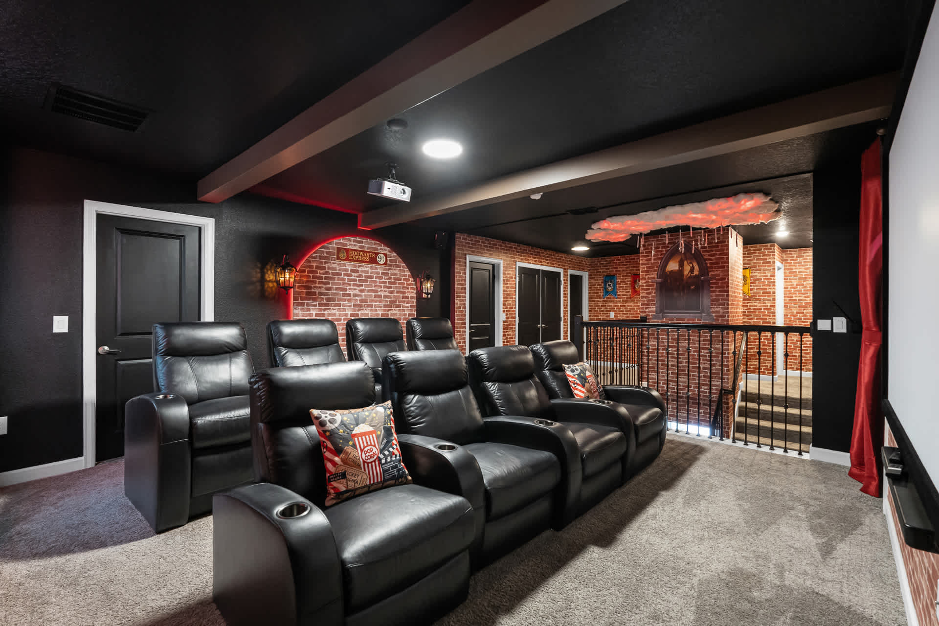 A home cinema room with multiple rows of black leather recliners, a large projection screen, and brick-patterned walls. A staircase with a black railing leads down into the room, and a glowing cloud decoration is visible.