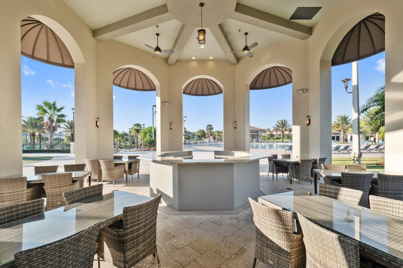 A spacious outdoor pavilion with arched openings, housing a central bar counter and multiple dining tables with wicker chairs. Palm trees and a large communal pool area are visible in the sunny background, creating a resort-like atmosphere.