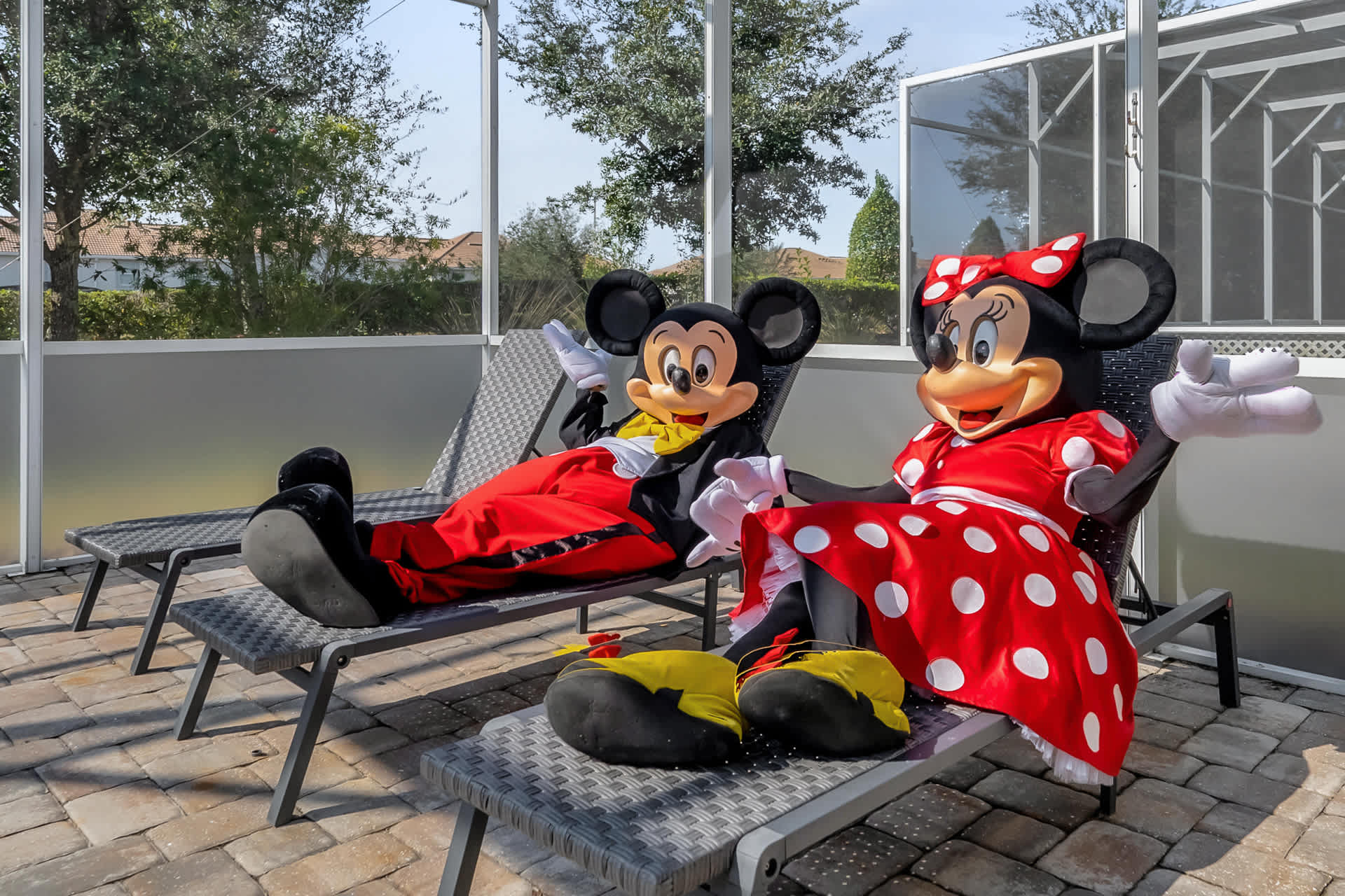 Mickey and Minnie Mouse figures relaxing on dark gray lounge chairs on a paved deck within a screened-in pool enclosure, with the private pool visible in the background.
