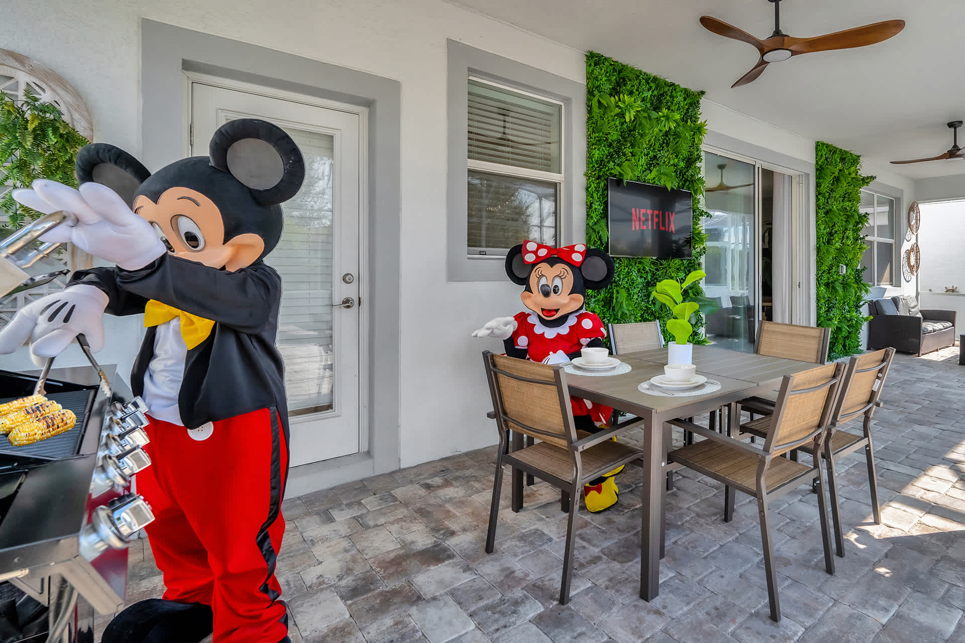 Covered outdoor patio featuring a dining table with chairs, a BBQ grill where Mickey Mouse is cooking, and Minnie Mouse seated at the table. The wall is adorned with artificial greenery and a "Netflix" sign.