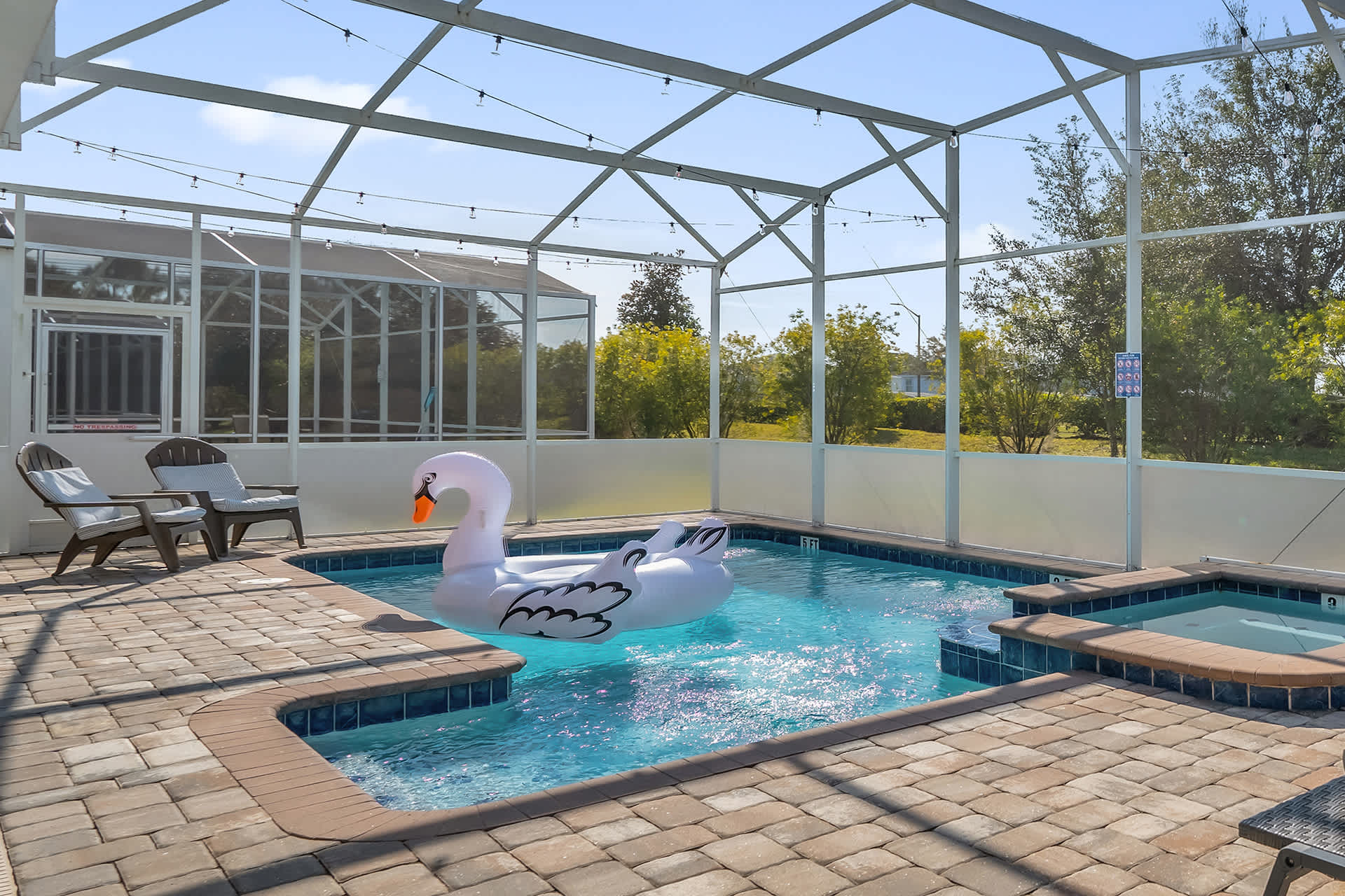 Screened-in private pool area featuring a rectangular swimming pool with a large white swan float, an adjacent hot tub, and two wooden lounge chairs on a paved deck, surrounded by a clear screen enclosure.