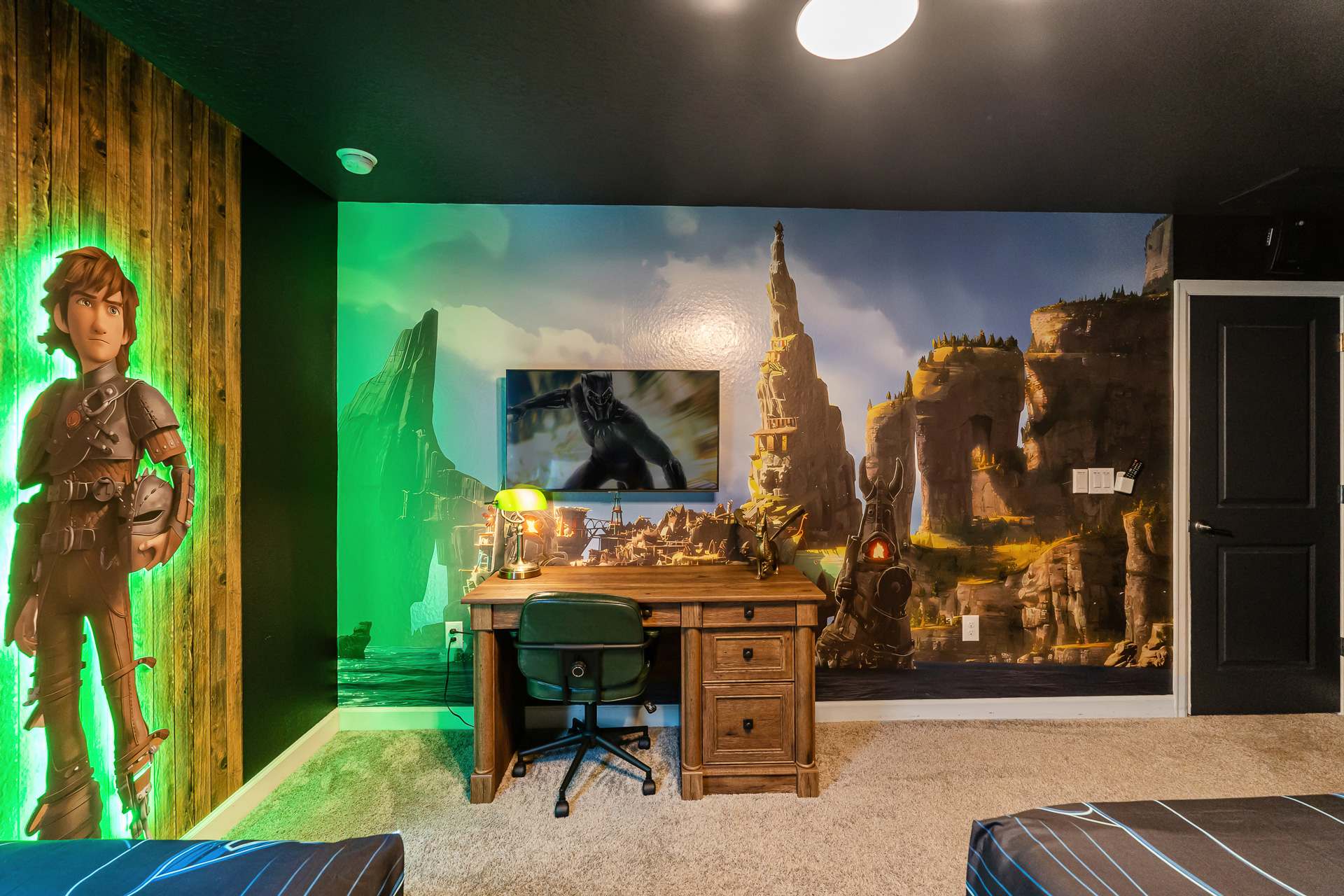 Themed bedroom featuring a large wall mural depicting a fantasy landscape, a wooden desk with a green chair, and a wall-mounted TV, with a character cutout on the left.