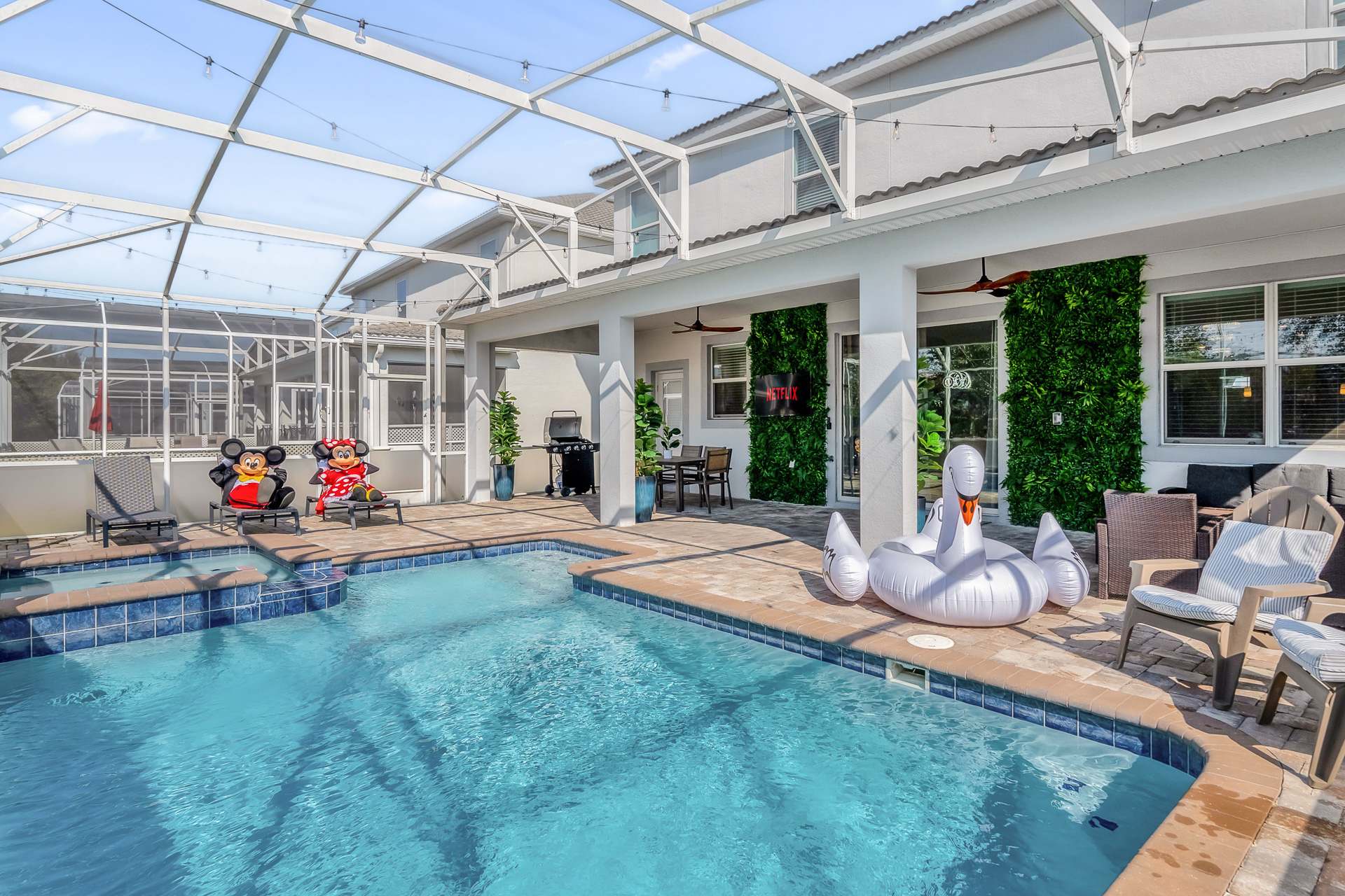 Bright daytime view of the screened-in private pool area, featuring a rectangular pool with a swan float, lounge chairs, a BBQ grill, and a paved patio with vertical garden accents.