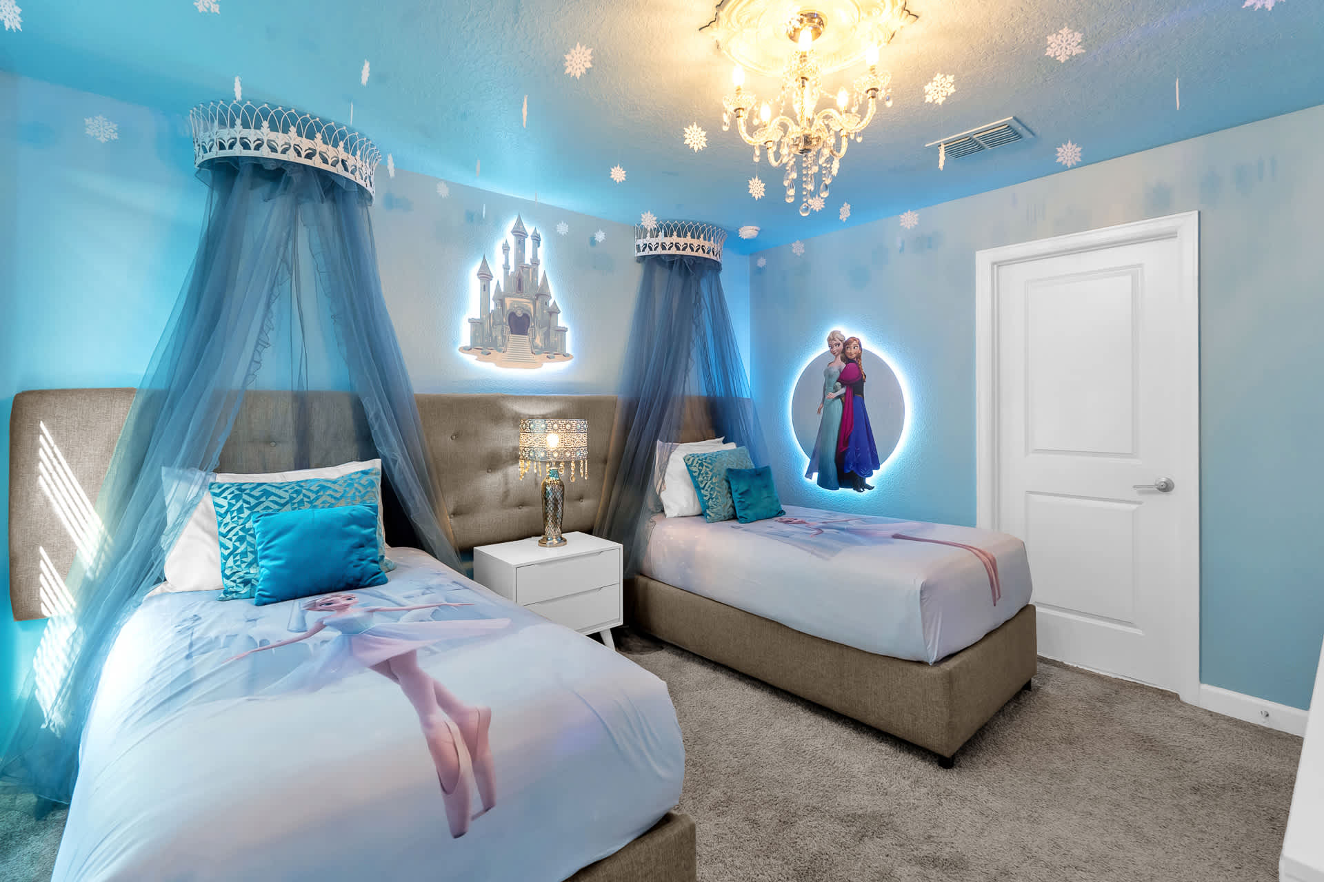 Themed "Frozen" bedroom with light blue walls, two twin beds with character bedding, canopy crowns, and illuminated wall art of a castle and characters.