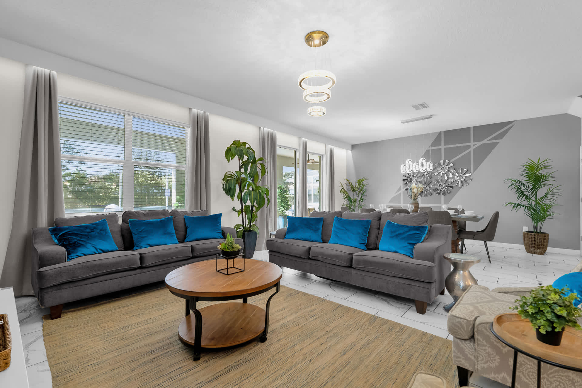 A bright and modern living room with two gray sofas adorned with blue pillows, a round wooden coffee table, and a large area rug. Large windows with blinds and curtains provide natural light, and a dining area is visible in the background.