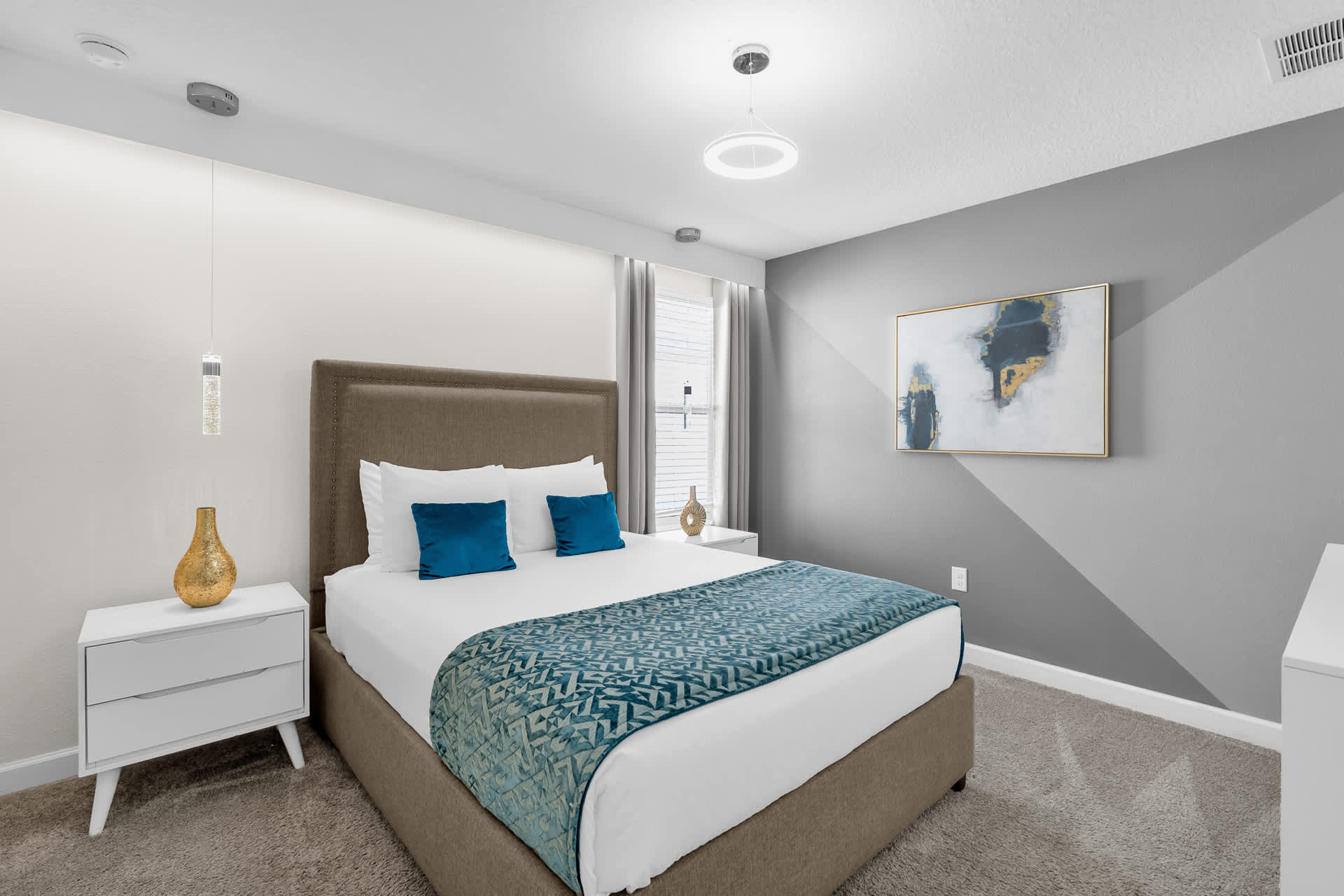 Modern bedroom with a gray upholstered queen bed, blue accent pillows, and a teal patterned throw. The room features a gray and white accent wall, a circular ceiling light, and two white nightstands with unique pendant lights.