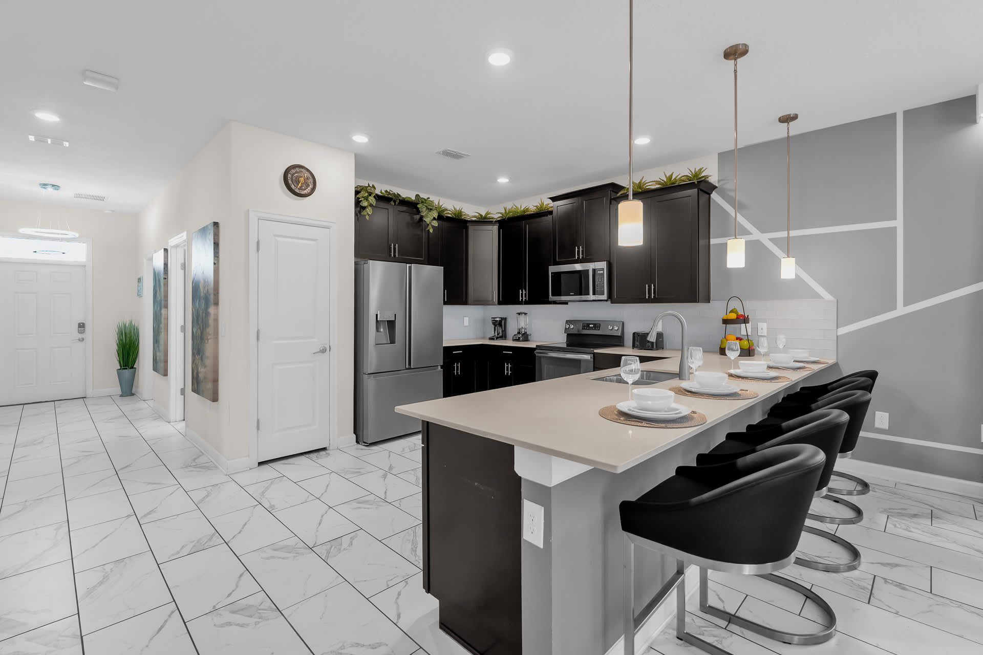 A contemporary kitchen featuring dark wood cabinets, light countertops, and a breakfast bar with four black bar stools. The entrance hallway with a white door and tiled floor is visible to the left.