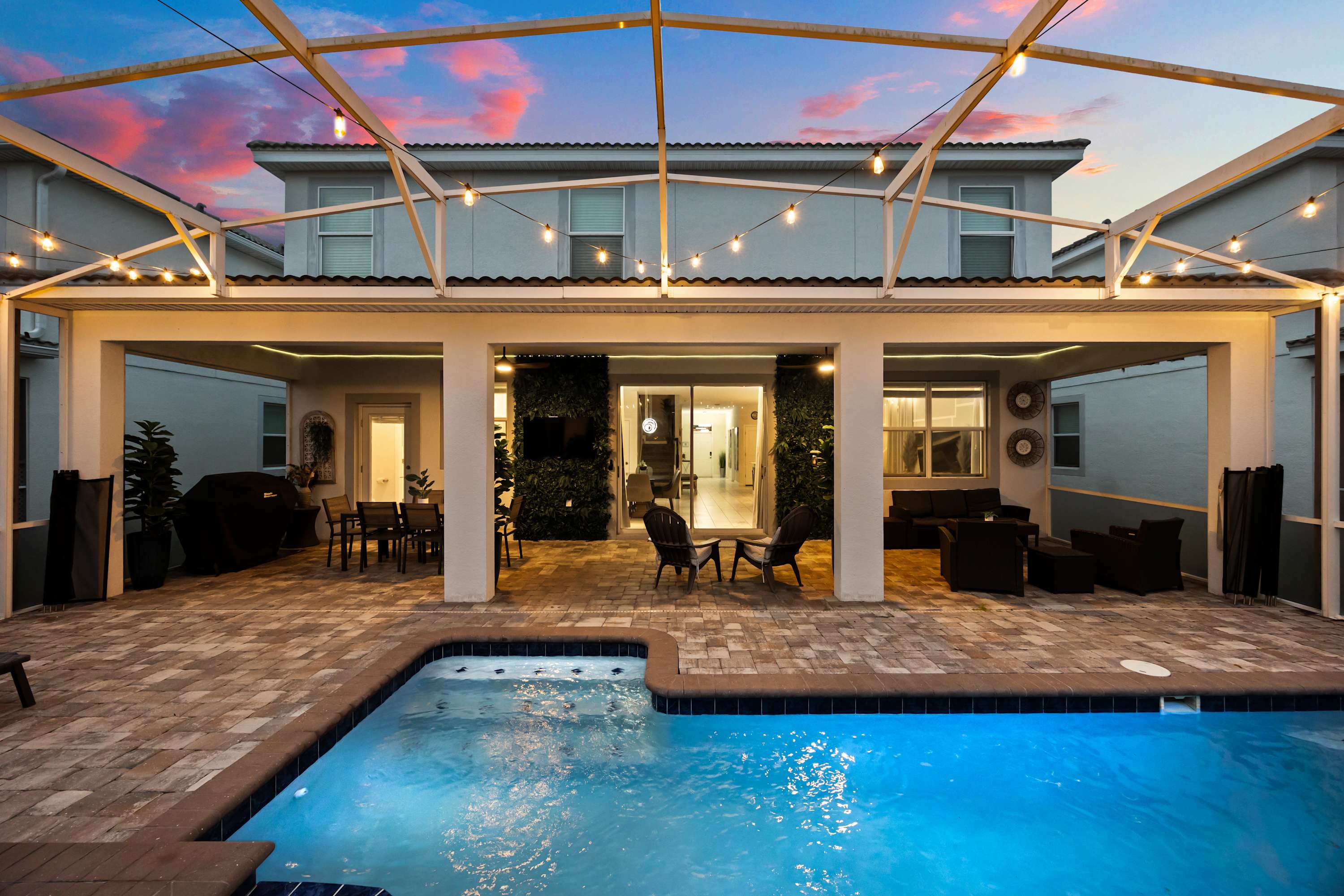 Expansive screened-in back patio with a private pool and hot tub, illuminated by string lights, set against a vibrant sunset sky and the two-story house exterior.