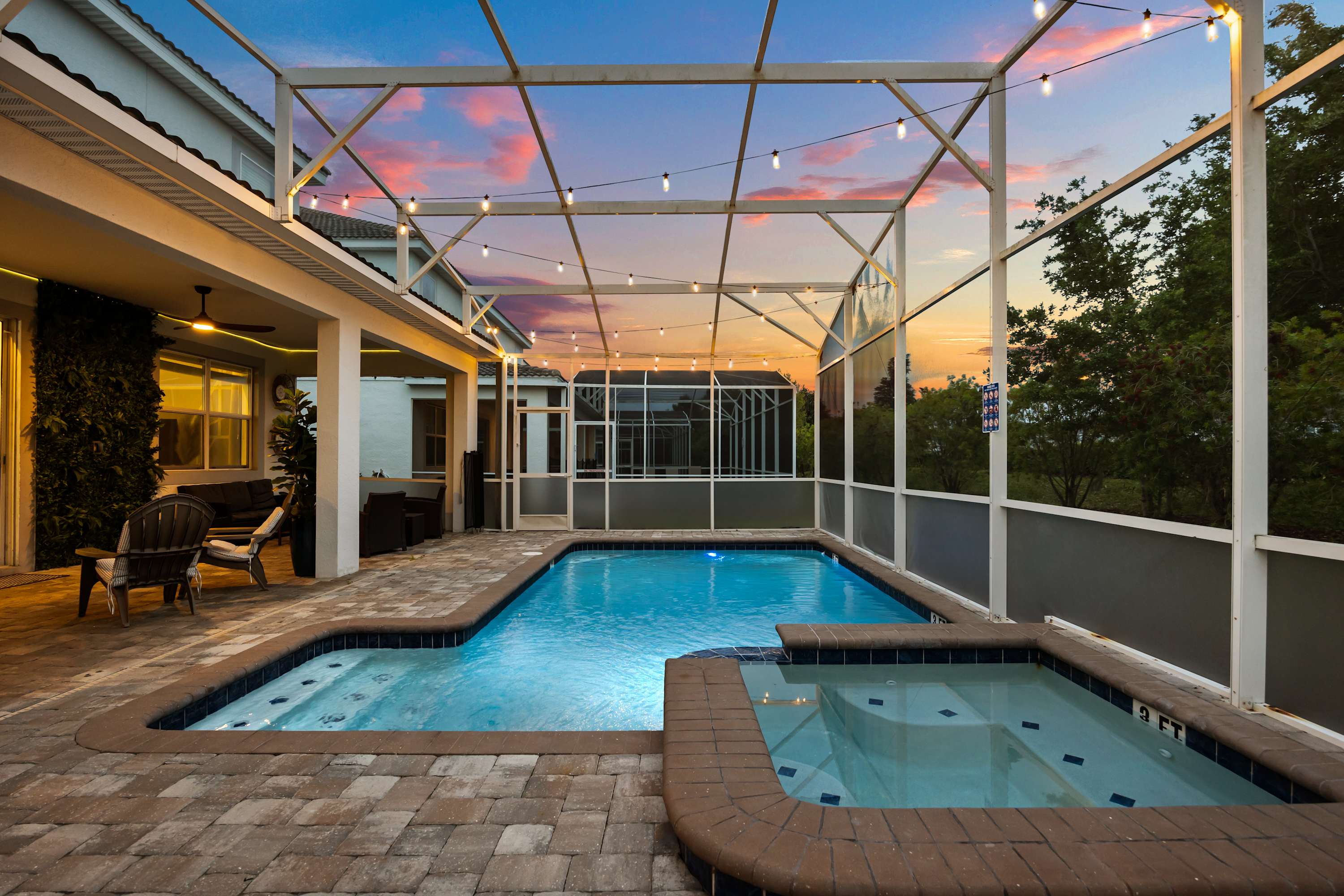 A large screened-in swimming pool and integrated hot tub are illuminated at dusk, surrounded by a paved patio and string lights overhead. The vibrant sunset sky is visible through the enclosure, enhancing the serene outdoor ambiance.