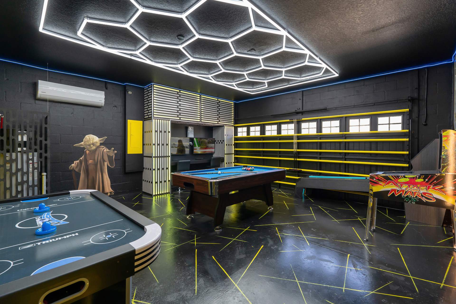 Expansive Star Wars-themed game room featuring an air hockey table, a pool table, and a pinball machine. The room has black walls with yellow accents, a Yoda statue, and a distinctive hexagonal LED ceiling light.