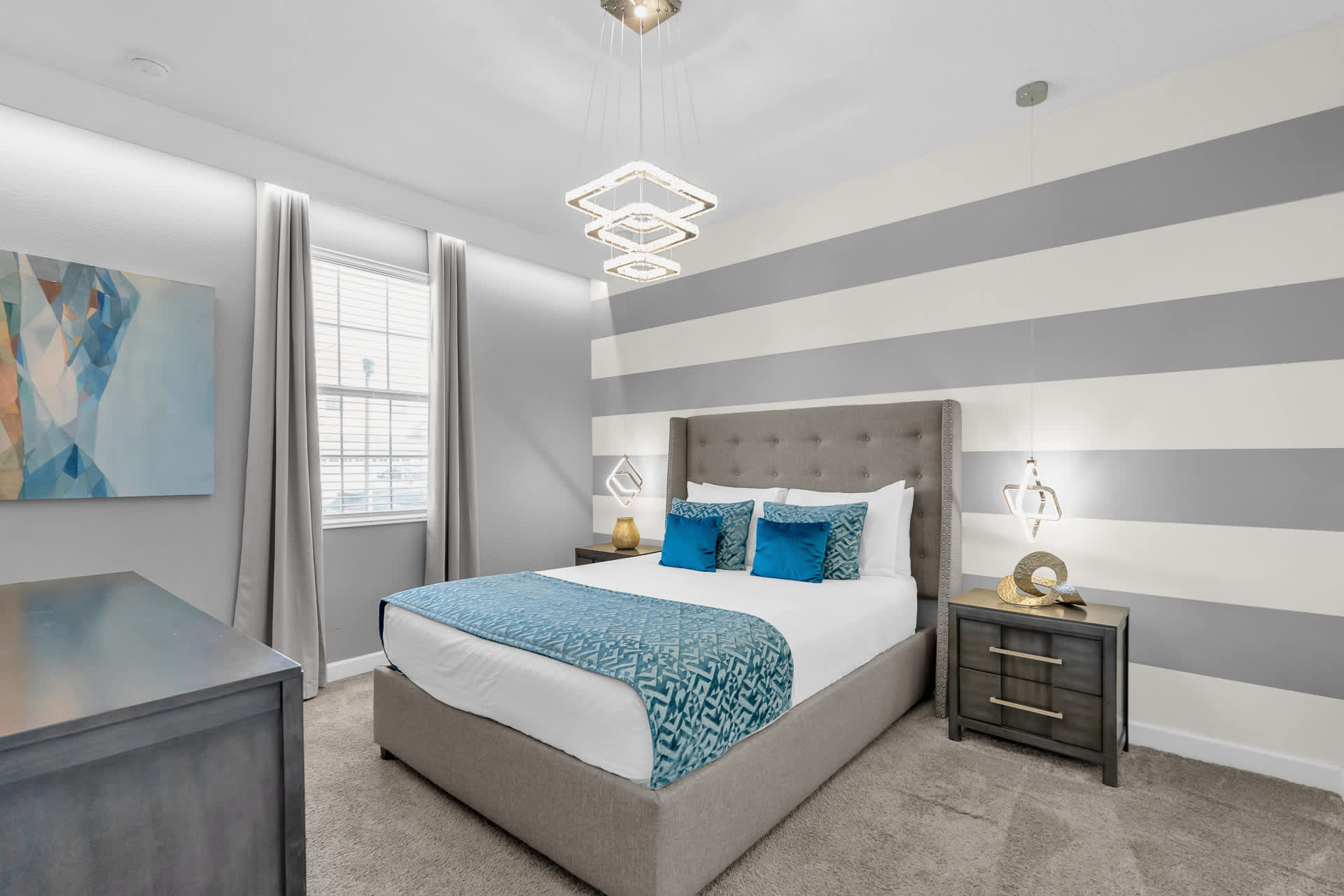 Contemporary bedroom featuring a gray upholstered king bed with blue accent pillows and a patterned throw. The walls are painted with horizontal gray and white stripes, complemented by a unique tiered square chandelier and two modern bedside lamps.