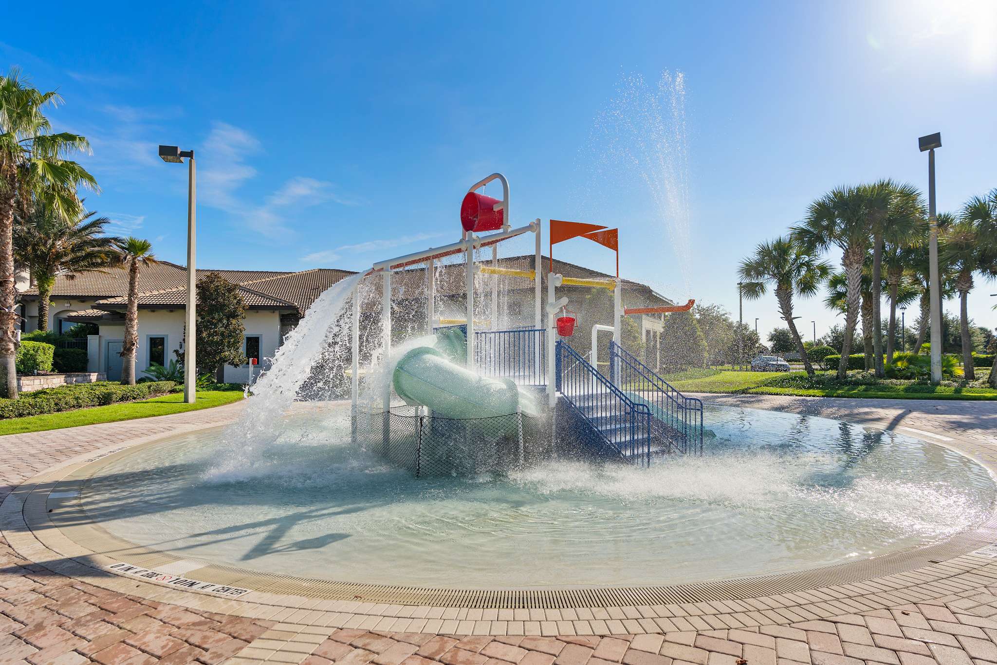 A vibrant children's splash pad features a large water play structure with a tipping bucket and a small slide, surrounded by shallow water and paved surfacing. Palm trees and resort buildings are visible in the background under a sunny sky.