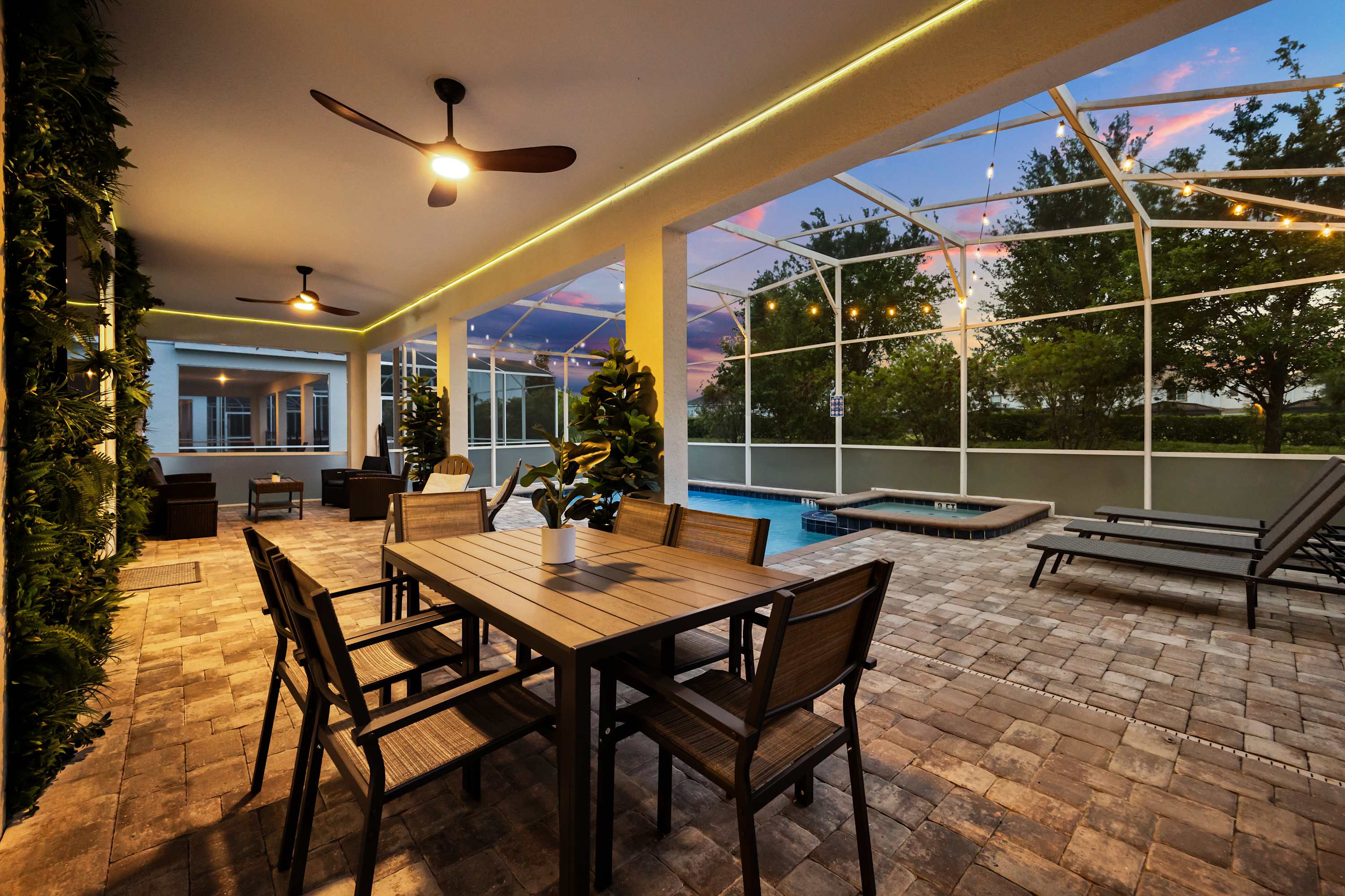 Spacious screened-in patio featuring an outdoor dining table, ceiling fans, and string lights, with a view of the private pool and hot tub under a colorful sunset sky.