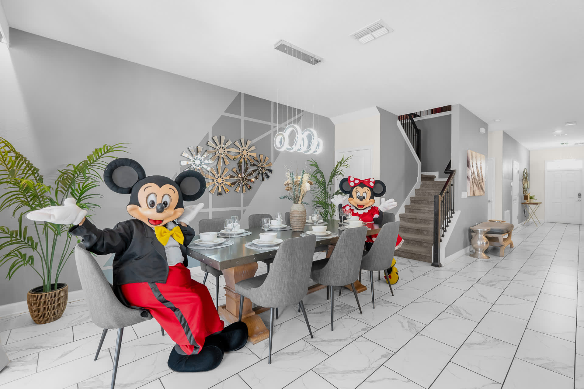 Modern dining room featuring a long wooden table with gray chairs, contemporary lighting, geometric wall decor, and two large Mickey and Minnie Mouse character figures.