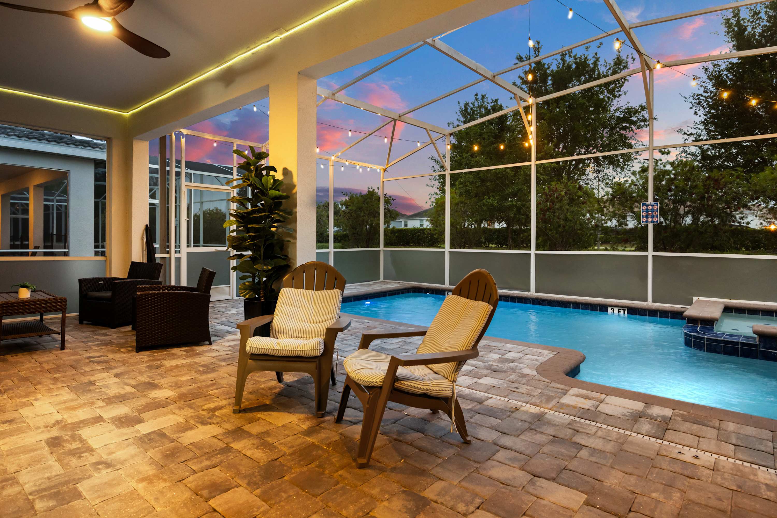 An inviting outdoor patio features two wooden Adirondack chairs with striped cushions, overlooking a screened-in swimming pool and hot tub under a string-lit enclosure at dusk. The paved patio extends to a covered area with additional seating.