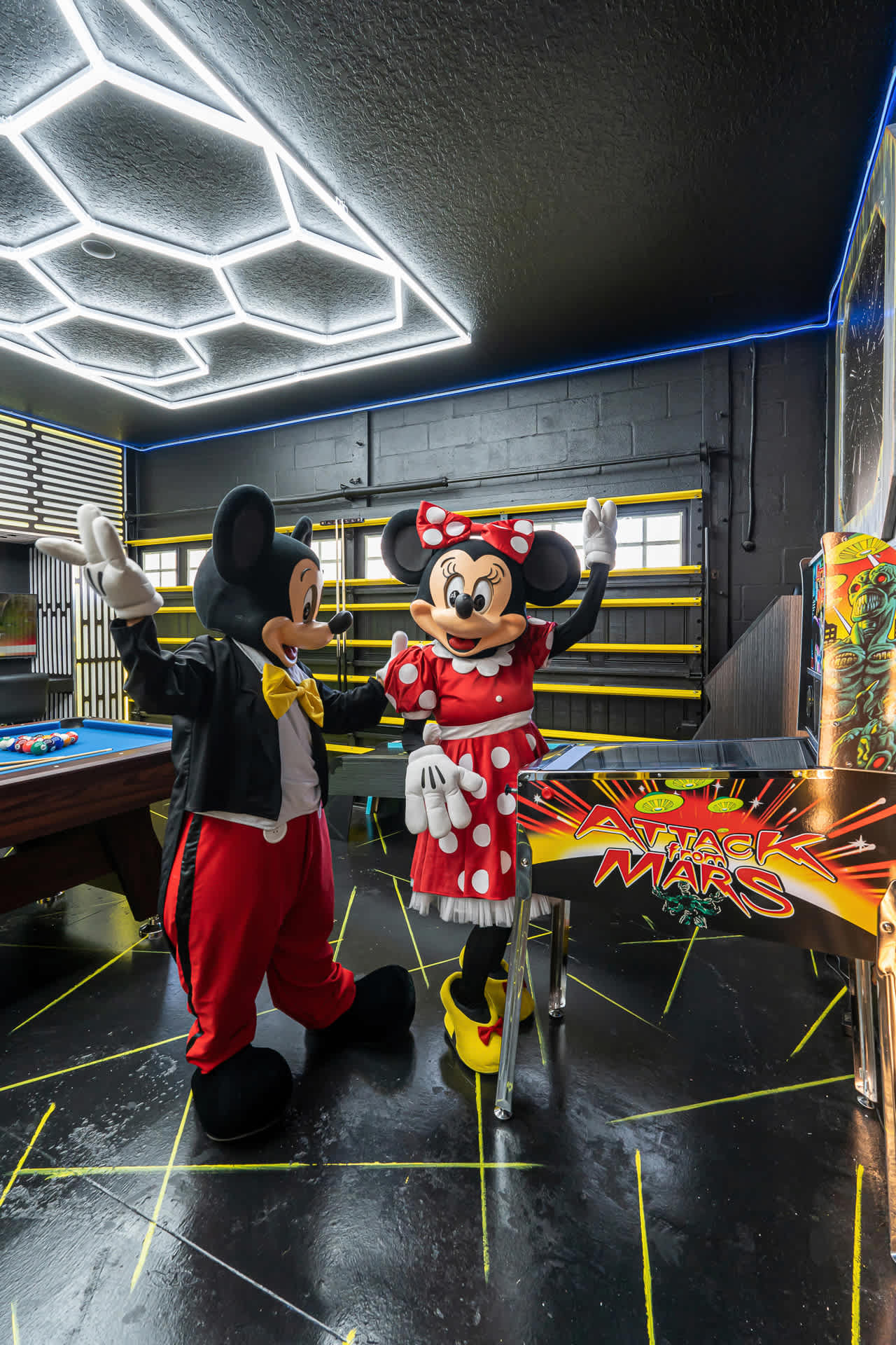 Mickey and Minnie Mouse figures standing next to a Star Wars-themed pinball machine in a game room with black walls, yellow floor lines, and a hexagonal LED ceiling light fixture.
