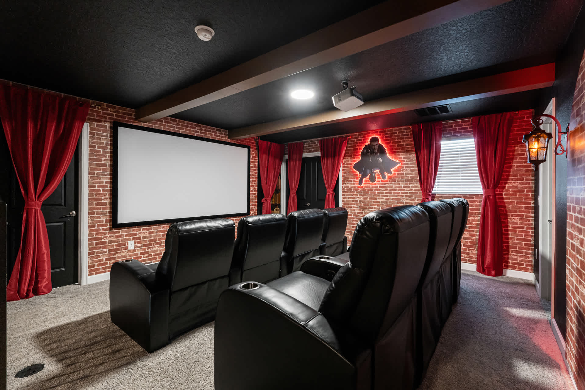 A home cinema room featuring black leather recliners, a large white projection screen, and brick-patterned walls with red curtains. A glowing red silhouette of a superhero is visible on the back wall.