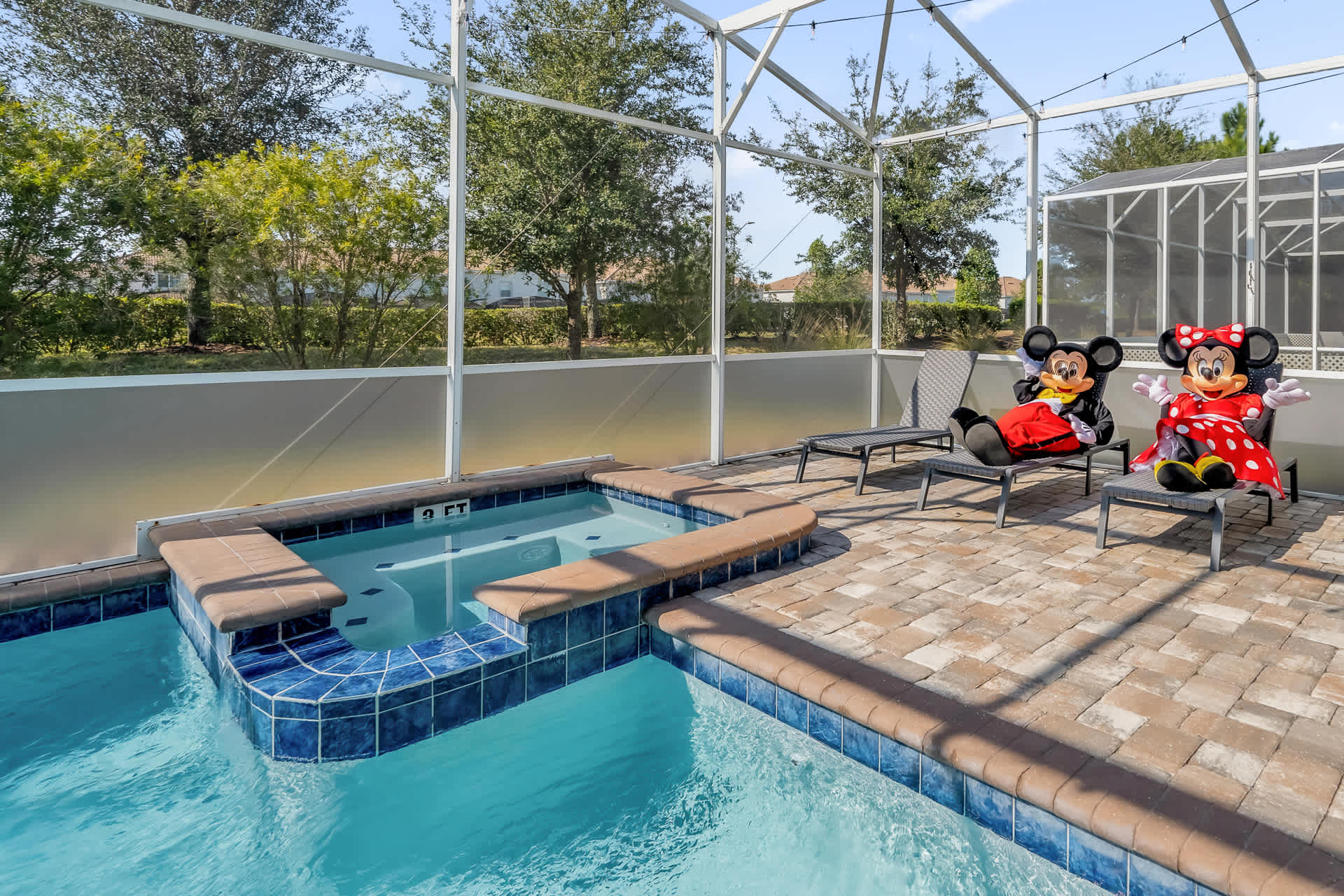 Screened-in private pool area with a hot tub in the foreground and two lounge chairs occupied by Mickey and Minnie Mouse figures, set on a paved deck with a view of trees beyond the enclosure.