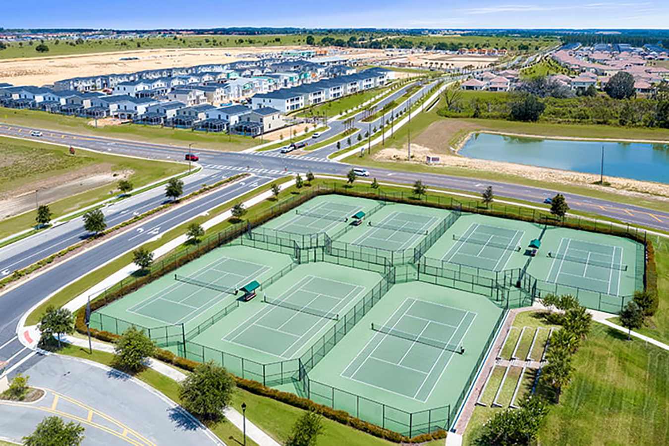 An aerial view showcasing a large complex of multiple green tennis courts, enclosed by fences, situated within a modern residential community. Roads, houses, and a tranquil pond are visible in the surrounding landscape.