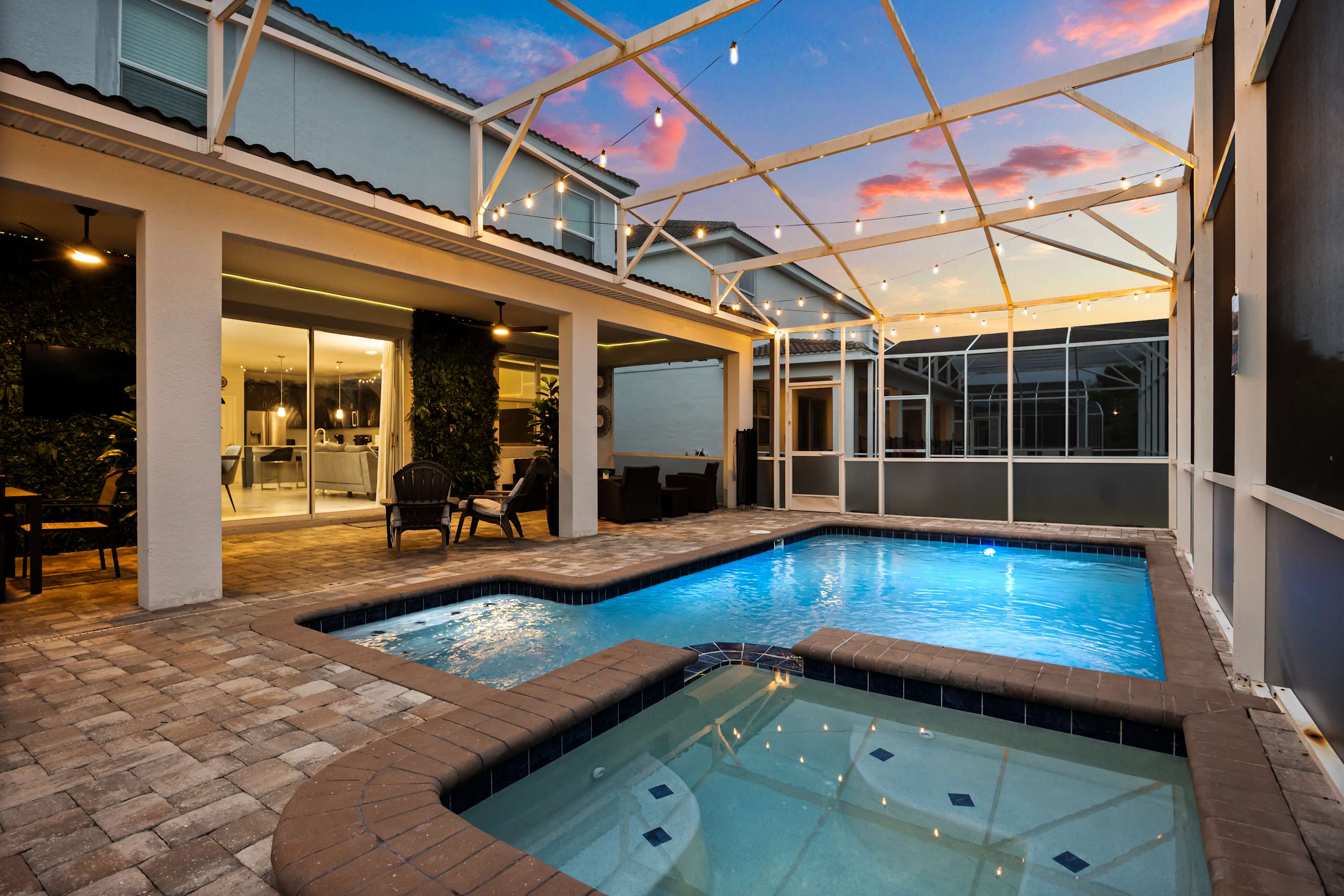 A spacious screened-in pool area features a large swimming pool and hot tub, bordered by a paved patio and the modern exterior of the house. String lights crisscross the enclosure, casting a warm glow as the sky transitions from day to night.