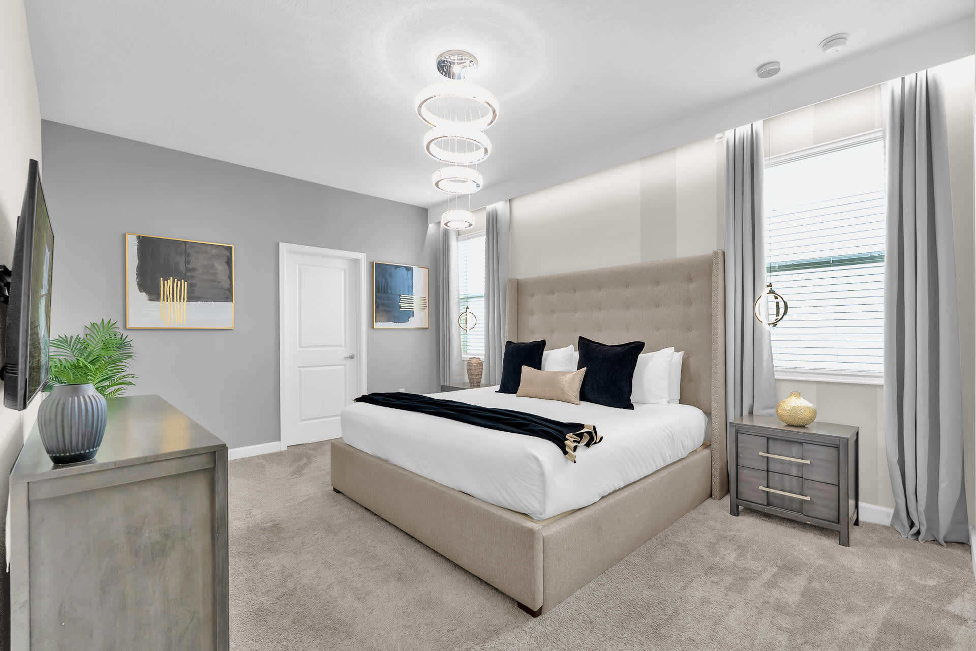 A stylish bedroom with a large upholstered king-size bed, two nightstands with lamps, and a dresser with a TV. The walls are painted in shades of gray and white, and large windows provide natural light.