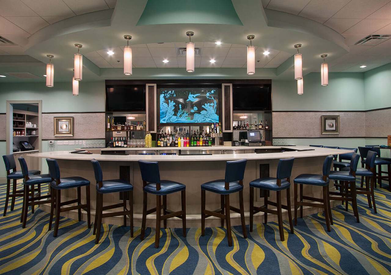 A stylish indoor bar with a curved counter and blue leather bar stools, featuring a large illuminated aquatic-themed display above the liquor shelves. The room has a vibrant blue and yellow wavy carpet and modern pendant lighting.