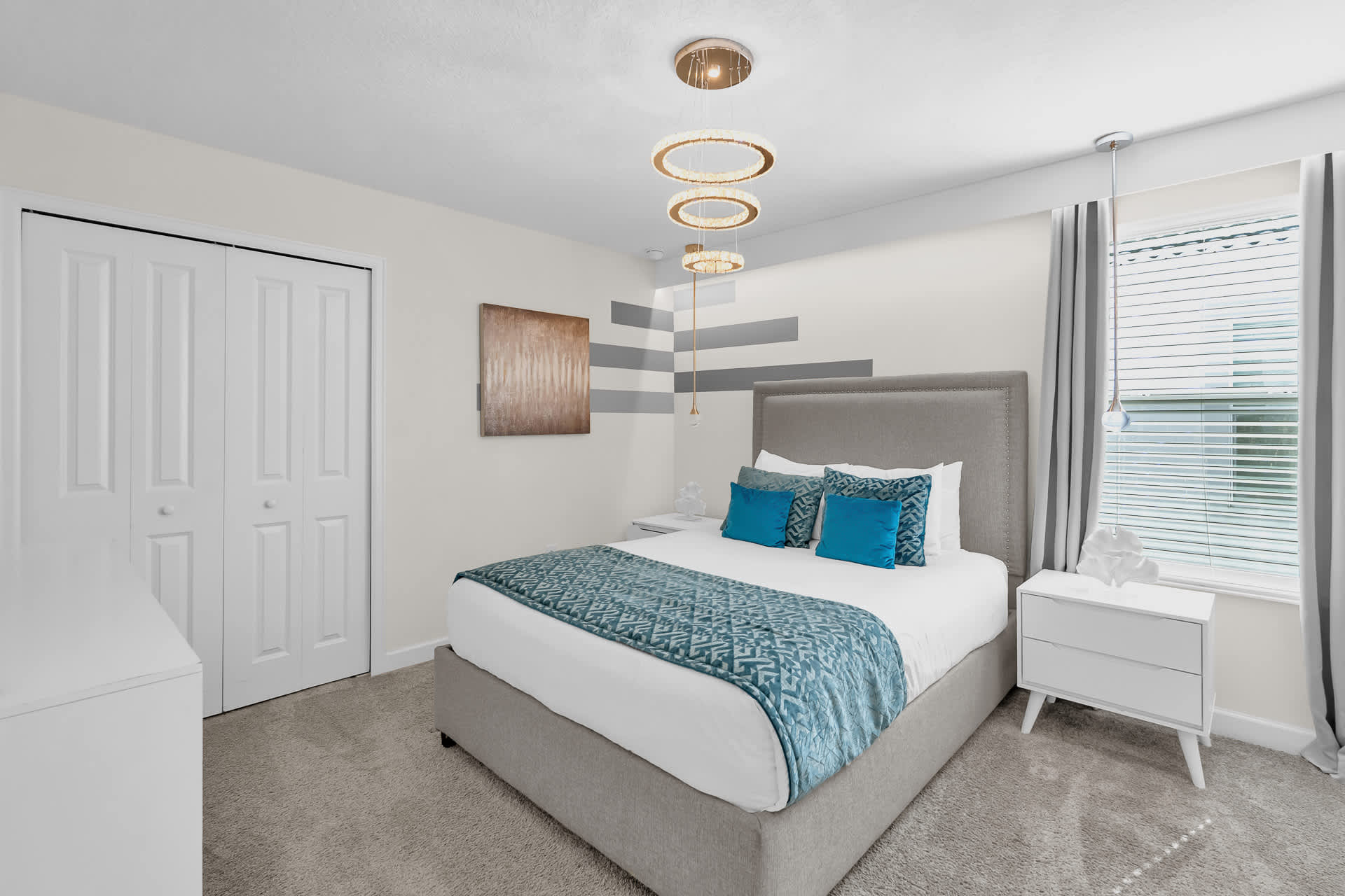 View of a modern bedroom showing a queen bed with a gray upholstered headboard, white nightstands, and a closet with bi-fold doors, illuminated by contemporary ring chandeliers.