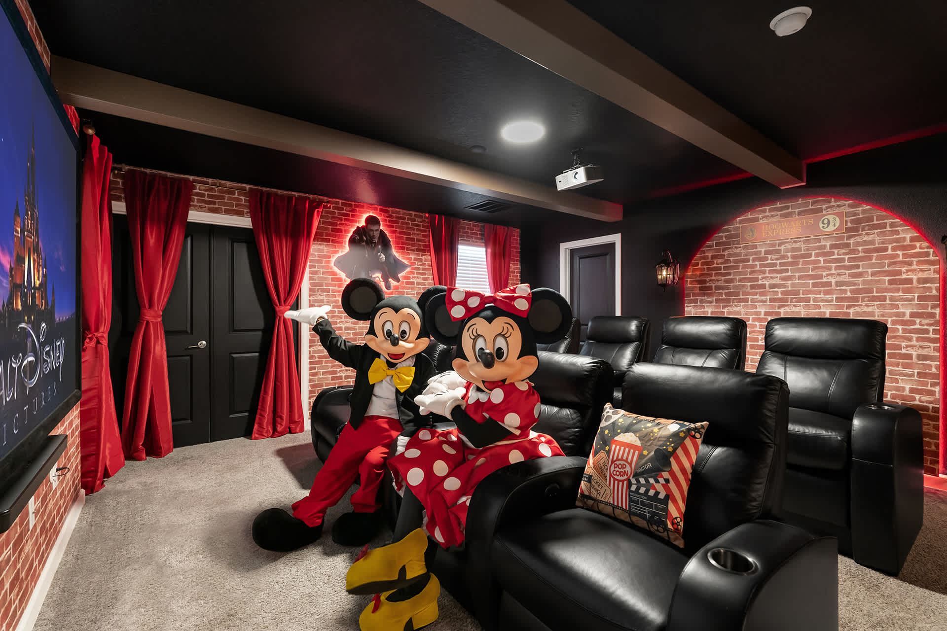 A themed home cinema room with black leather recliners, a large projection screen displaying a Disney castle, and brick-patterned walls with red curtains. Mickey and Minnie Mouse characters are seated in the front row.