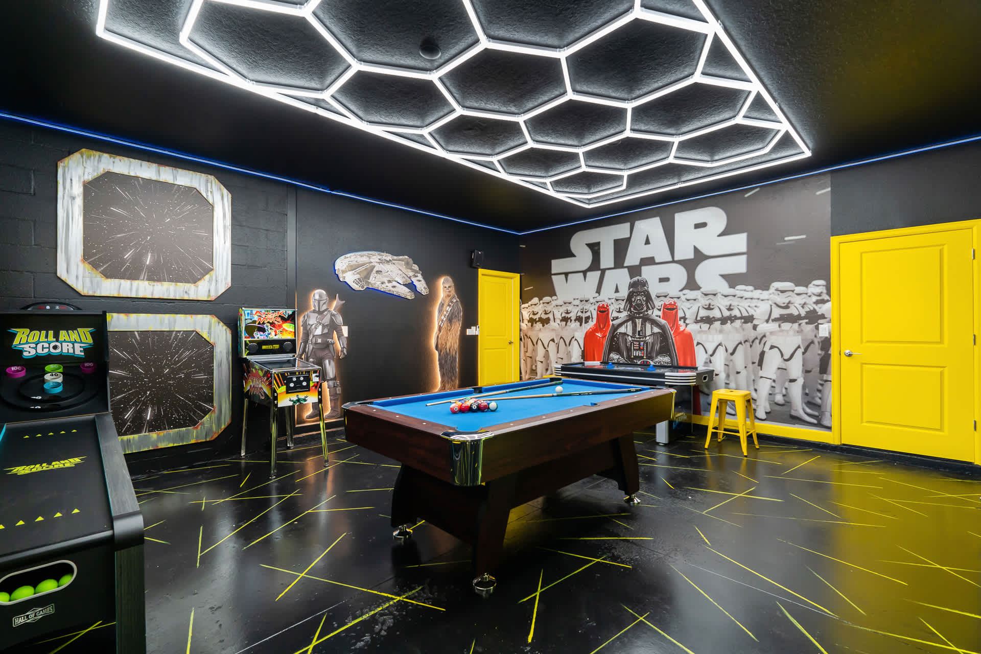 Star Wars-themed game room featuring a pool table in the center, a pinball machine, and an arcade game. The room has black walls with yellow accents, a black floor with yellow lines, and a striking hexagonal LED ceiling light fixture.