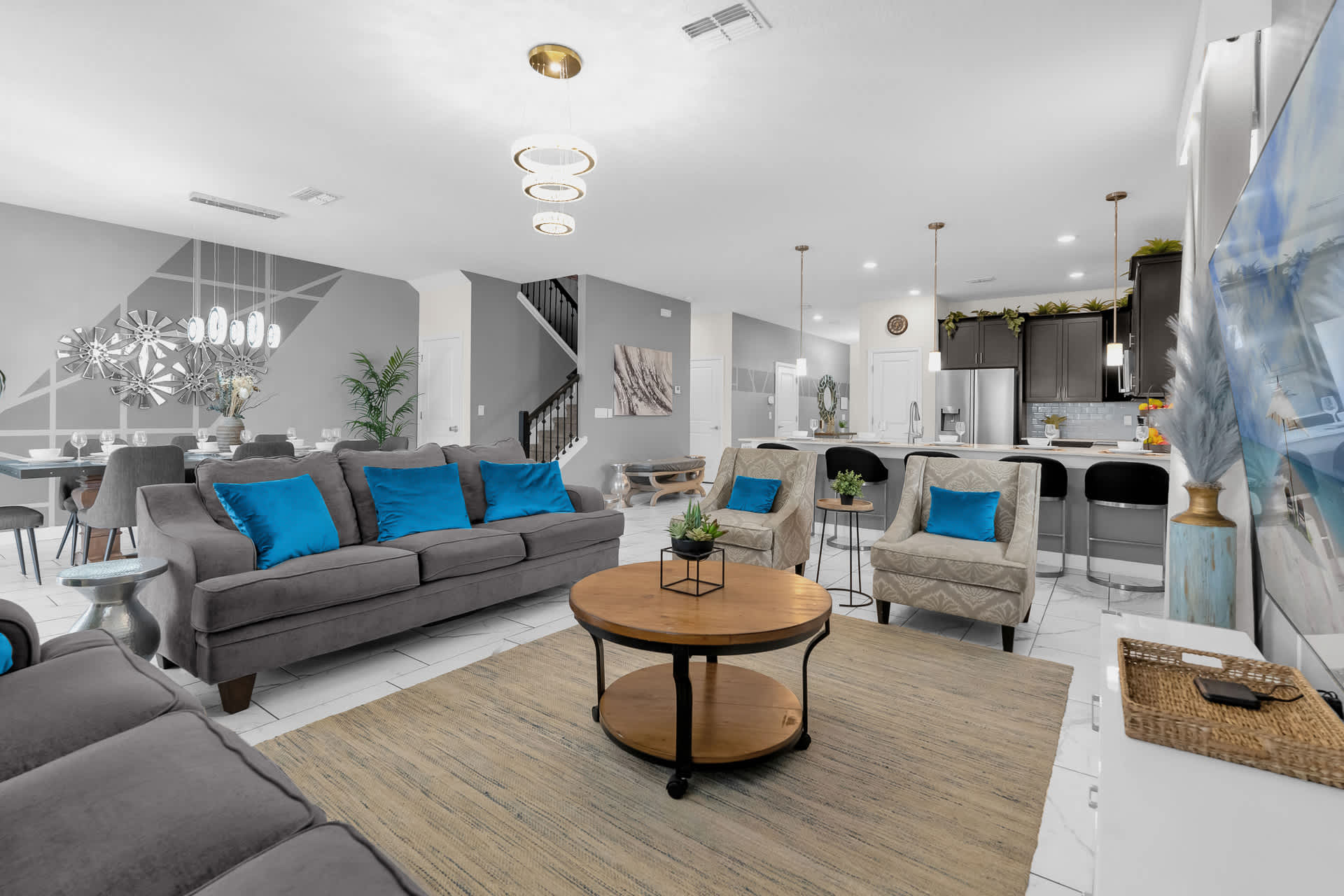 An open-concept living area with a gray sofa, two armchairs, and a round wooden coffee table on a large rug. The dining area and kitchen with a breakfast bar are visible in the background.