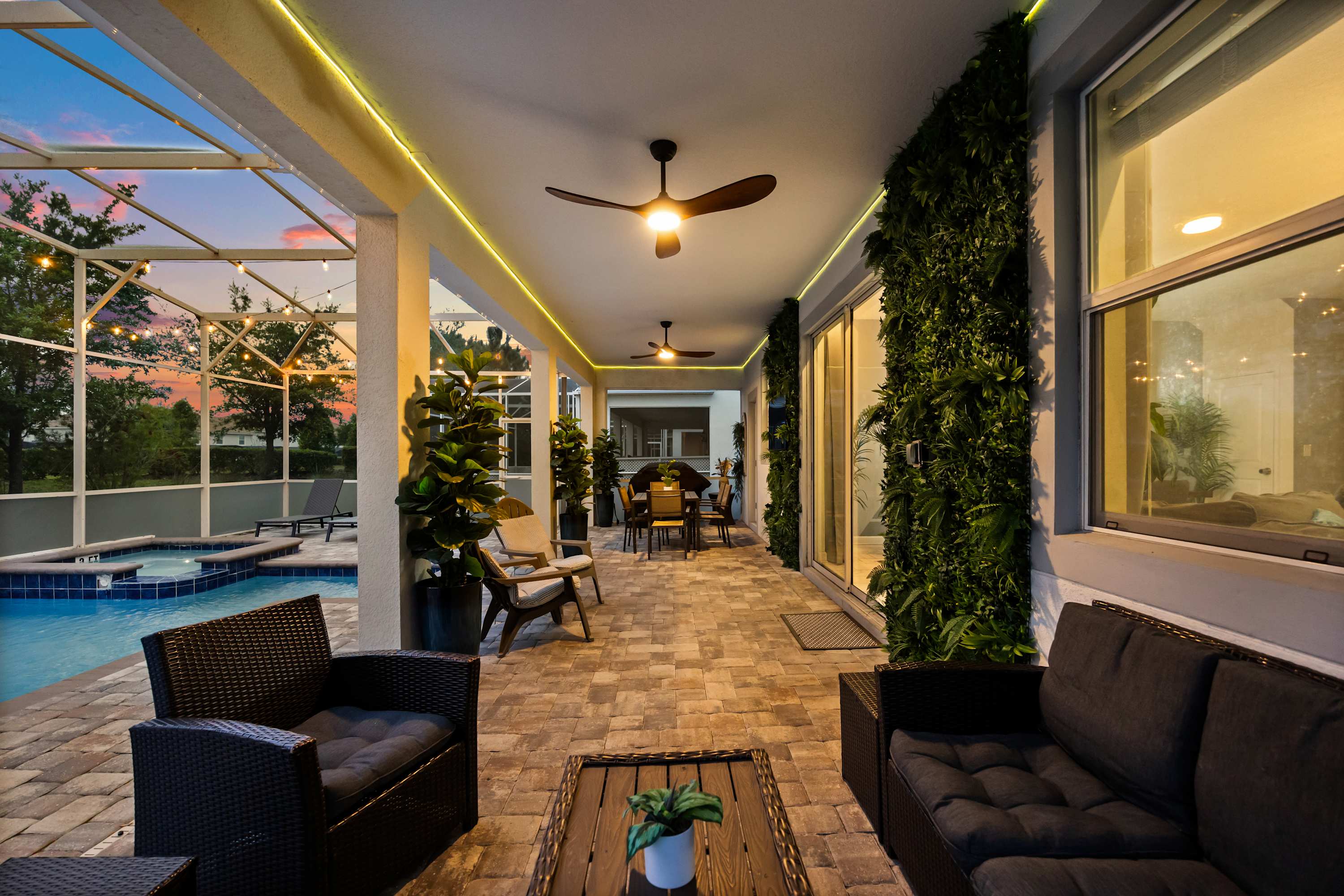 A long, covered outdoor patio features multiple seating areas, including dark wicker sofas and chairs with cushions, and a dining table, all illuminated by ceiling fans and strip lighting. A lush vertical garden adorns the wall, overlooking the screened pool area.