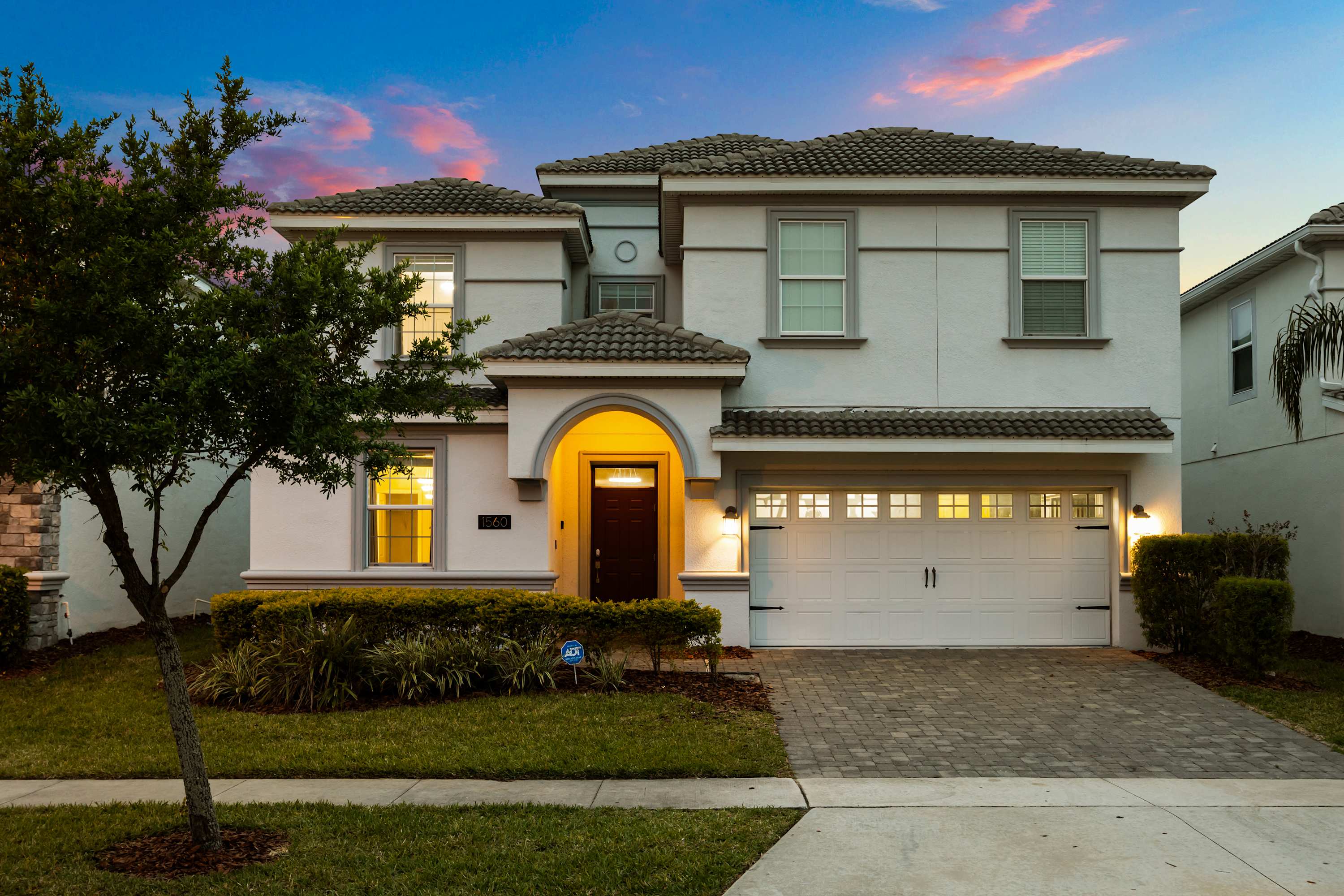 Modern two-story house exterior with a light gray facade, a paved driveway leading to a two-car garage, and an arched entryway, illuminated at dusk.