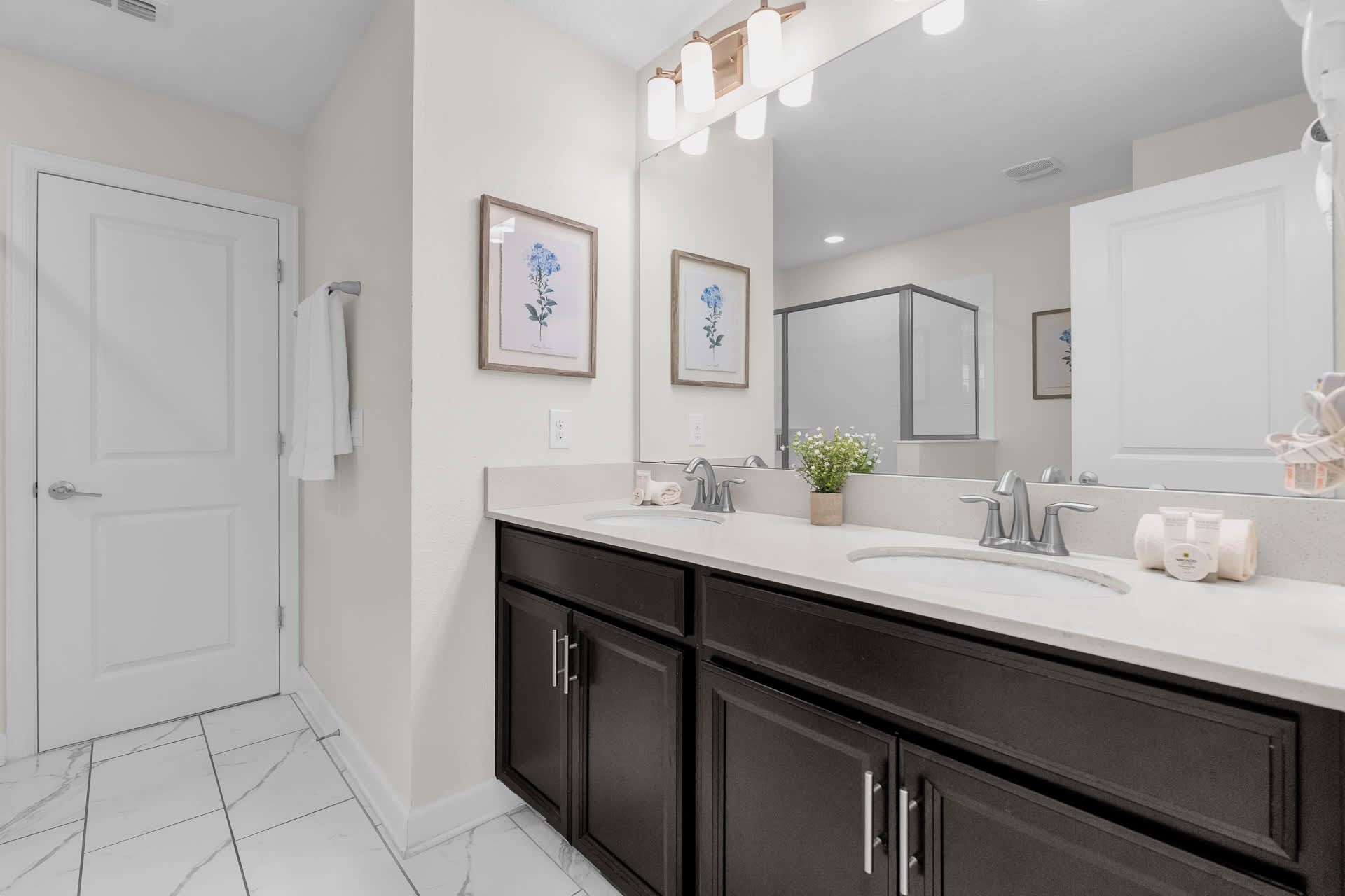 Modern bathroom with a double vanity featuring dark wood cabinets, light quartz countertops, and two rectangular sinks. The room has white tiled floors and a large mirror reflecting a shower and a white door.